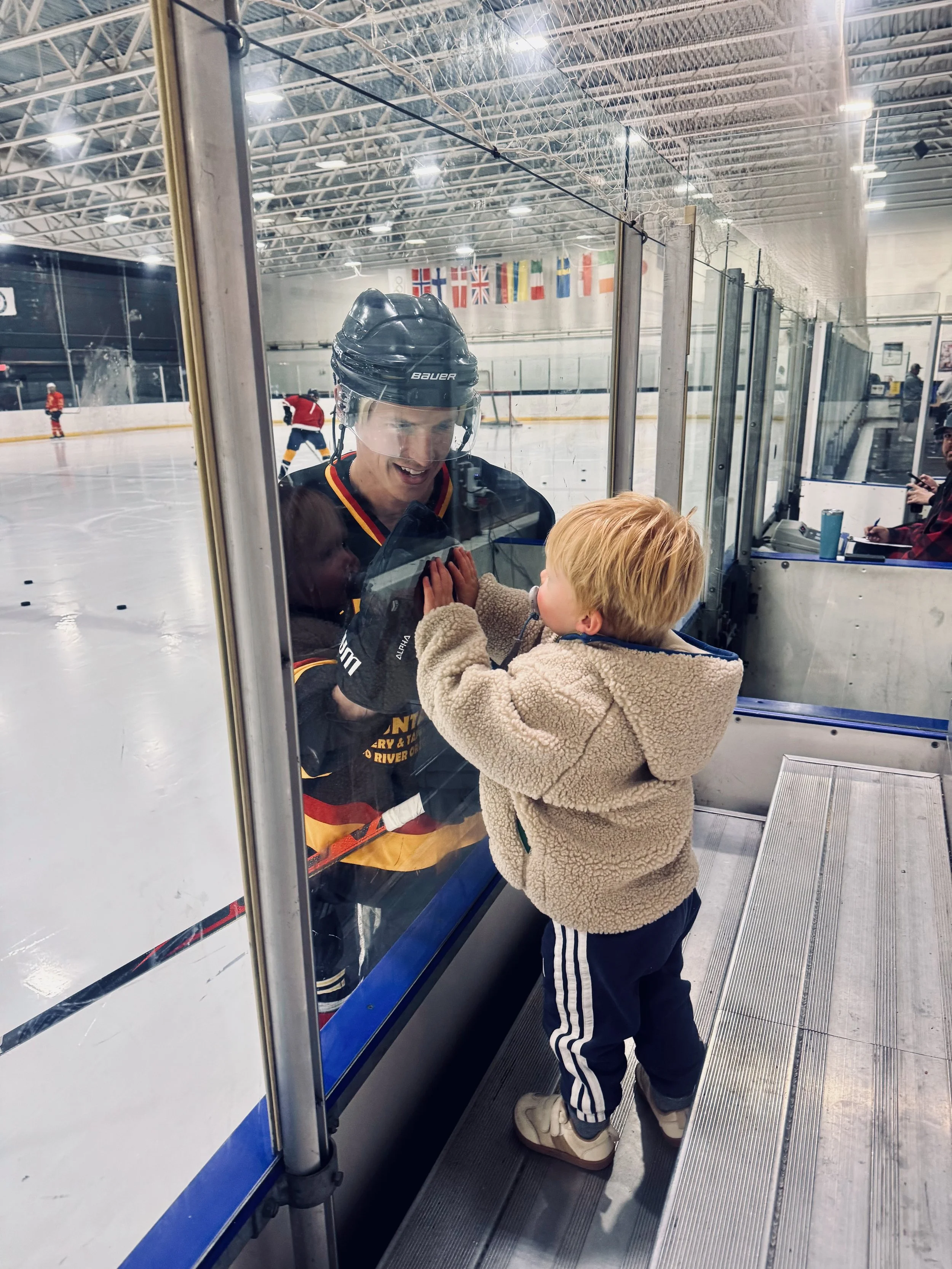 A young boy stands at the glass barrier watching a hockey player, who is smiling and looking at the boy, in an indoor ice rink. Several players are skating in the background, with flags hanging from the ceiling.