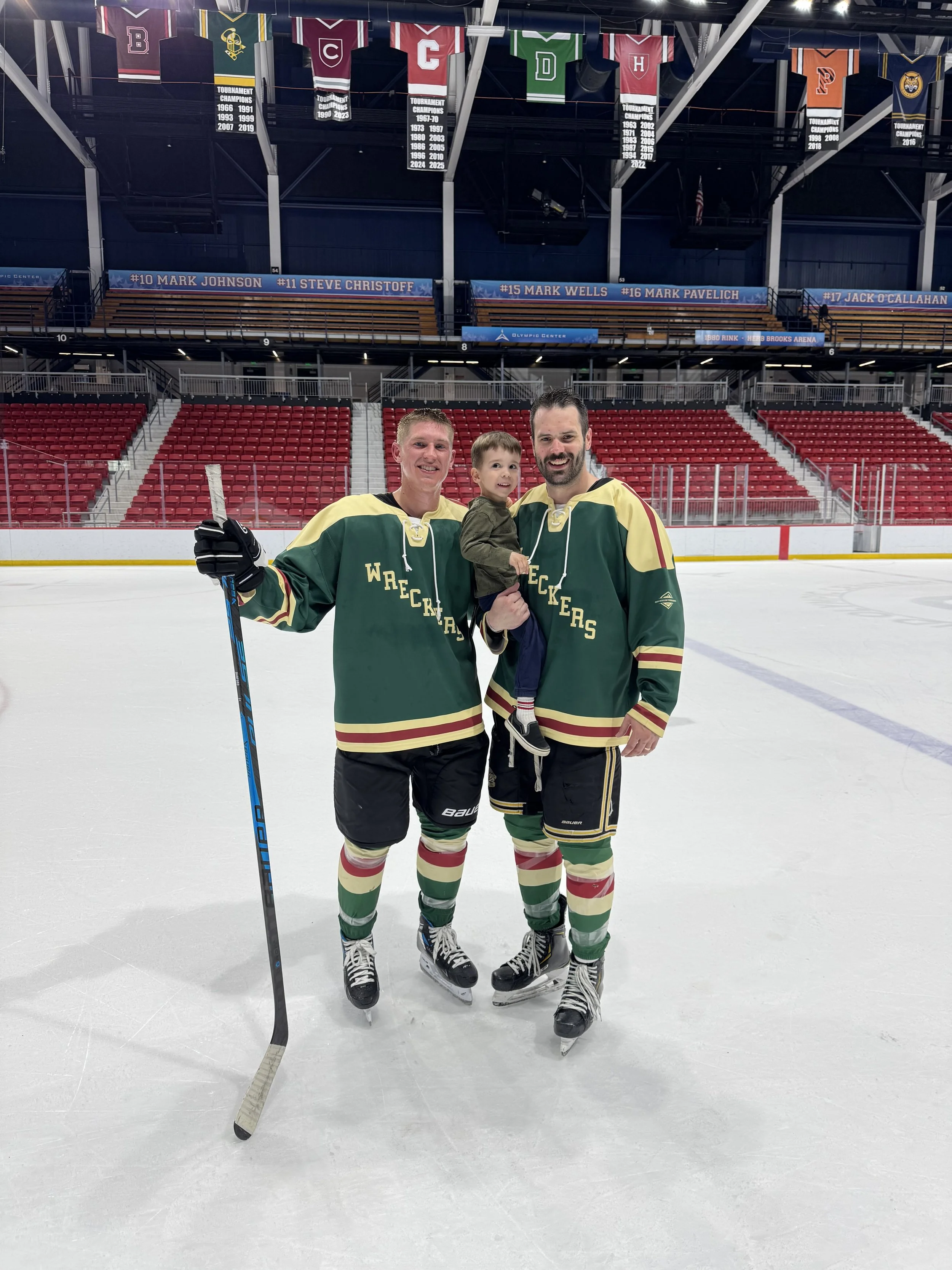 Two hockey players and a young boy on an ice rink, all wearing green jerseys with yellow and red stripes, smiling at the camera. The players are holding hockey sticks, and the rink is empty with red seats and banners hanging above.