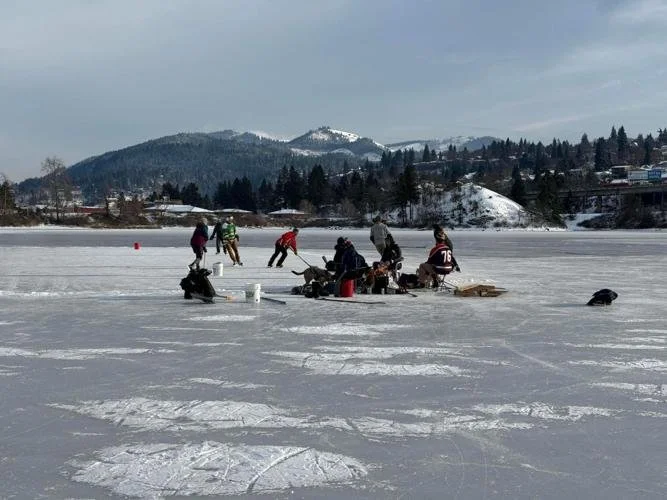 People ice fishing on a frozen lake with snow-covered mountains and trees in the background.