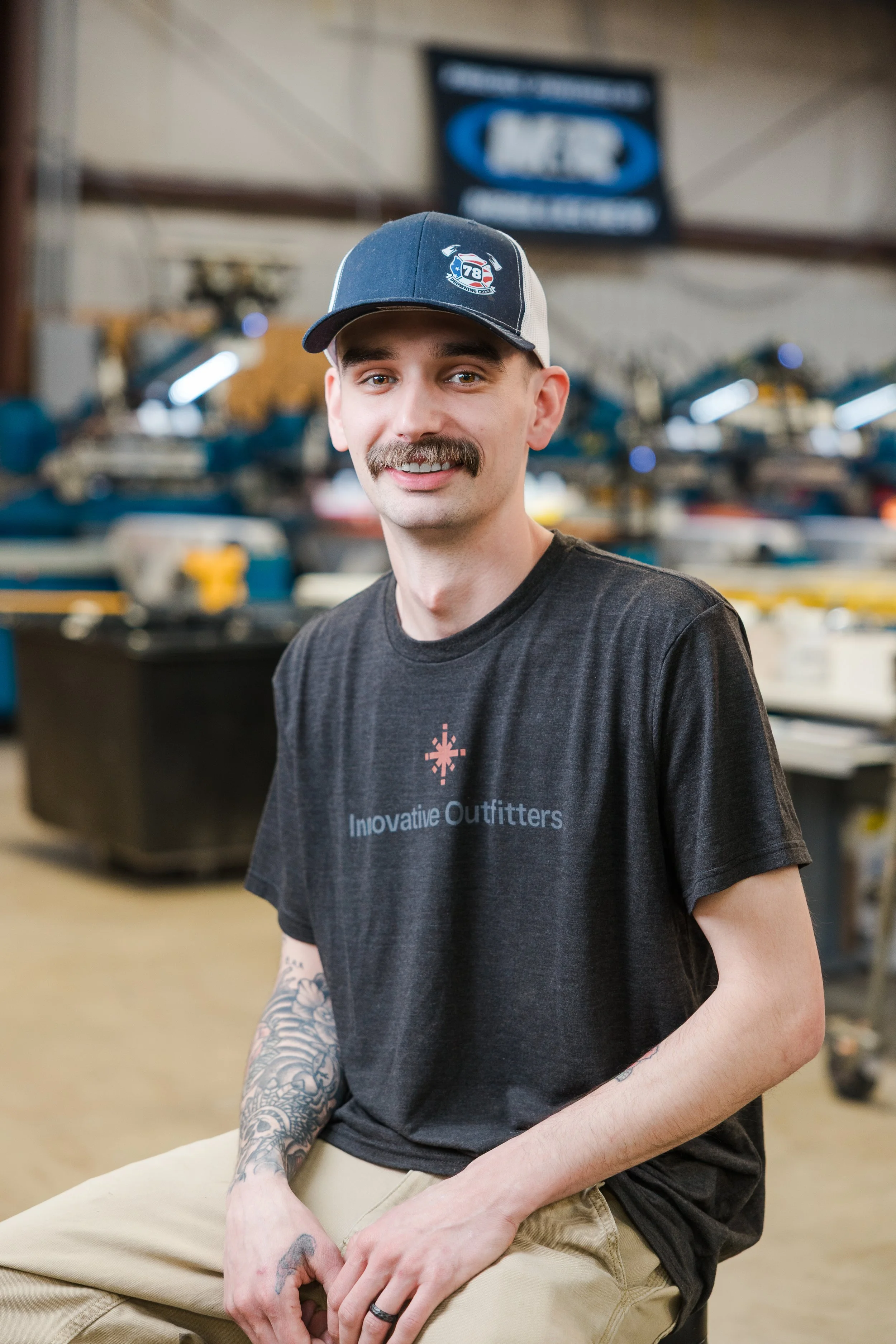 A young man with a mustache and tattoos on his arm is sitting in a workshop, wearing a black 'Innovative Outfitters' T-shirt and a blue trucker hat, with tools and equipment in the background.