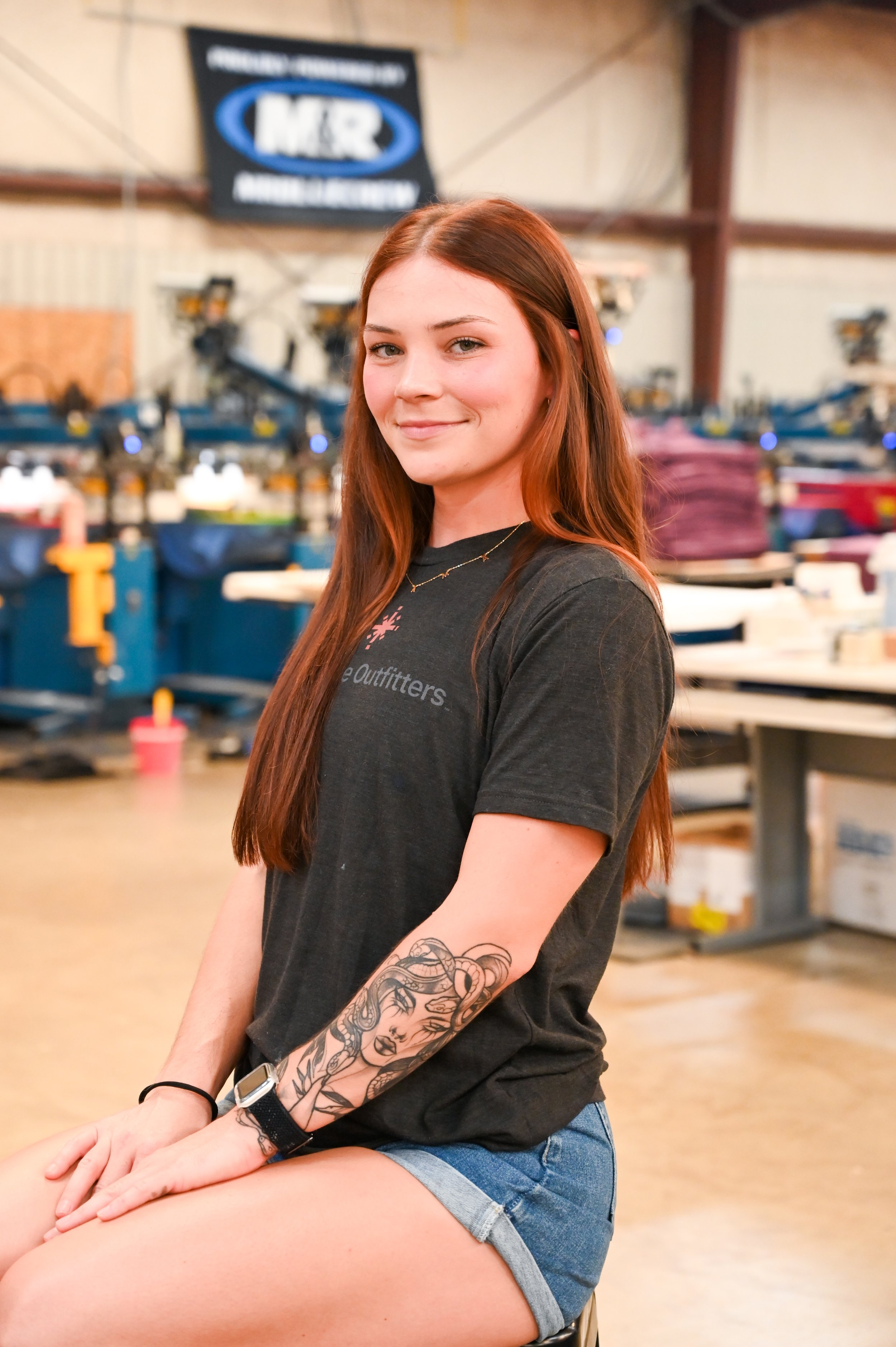 A young woman with long red hair, smiling, sitting in a warehouse or factory setting with machines and boxes around her.