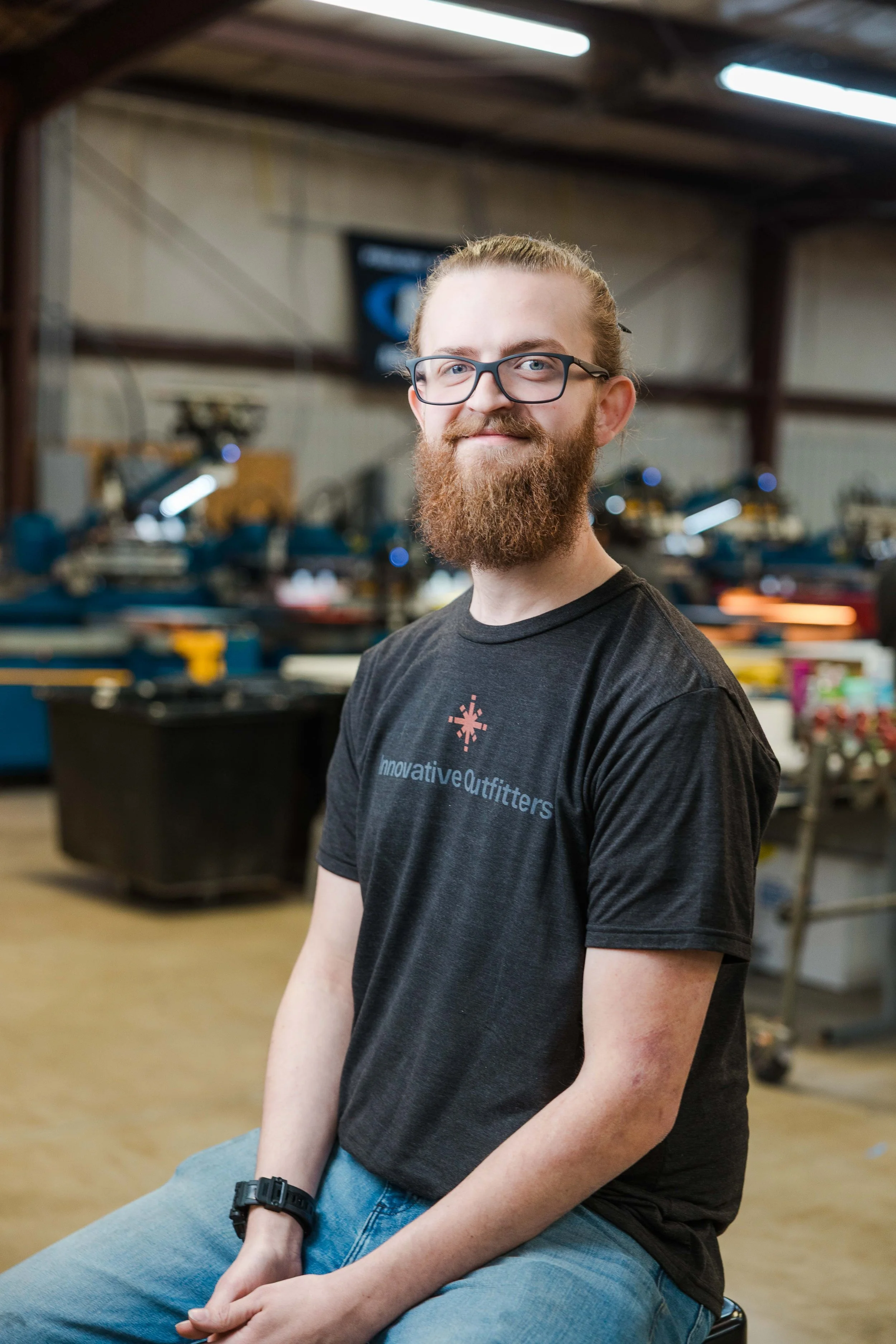 A young man with glasses and a beard sitting in a workshop or industrial space, wearing a black t-shirt with a logo and the words 'innovative Outfitters.'