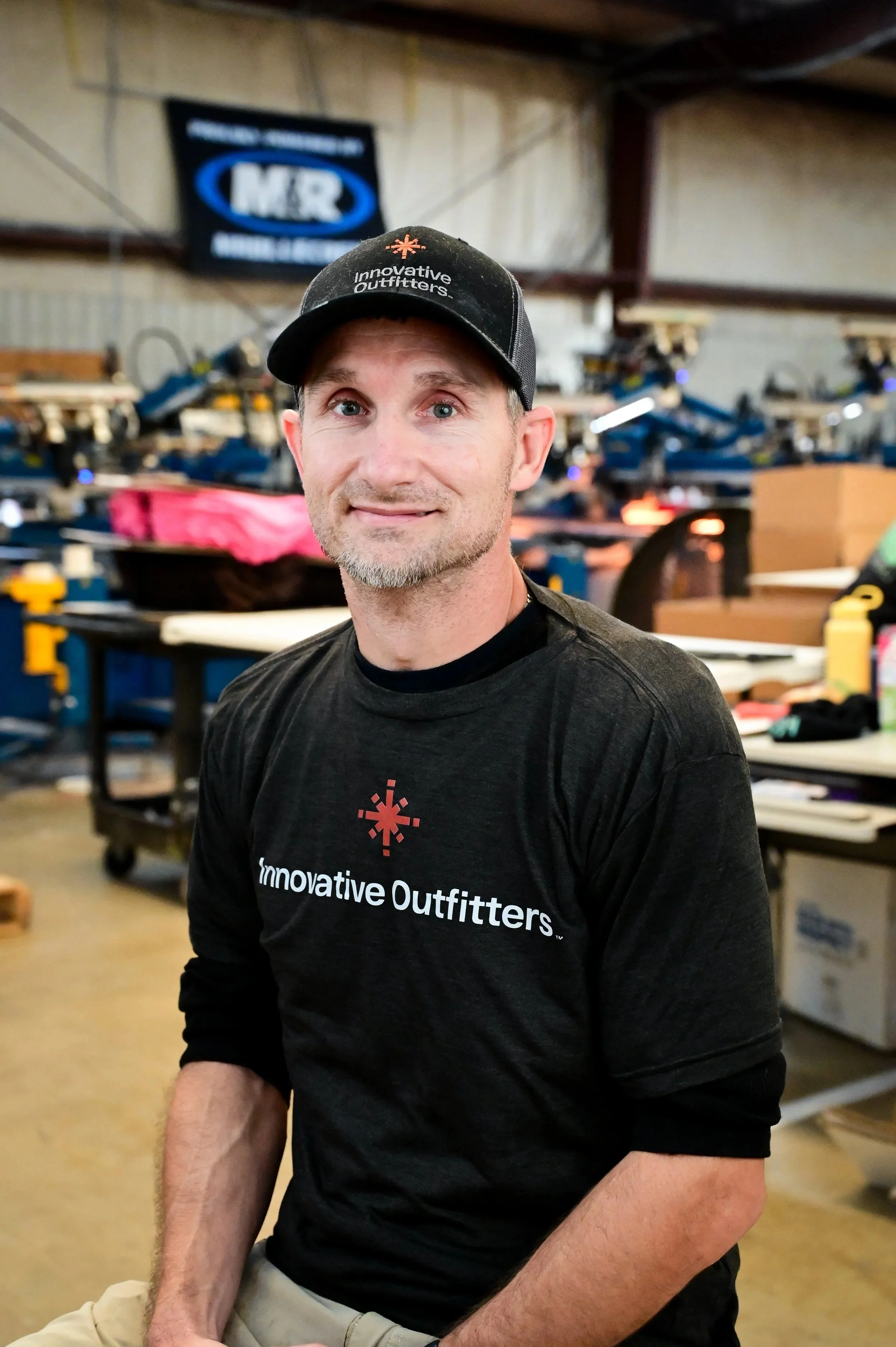 A man wearing a gray polo shirt and a black cap with the words 'Innovative Outfitters' stands in a warehouse or manufacturing facility with various equipment and tools in the background.