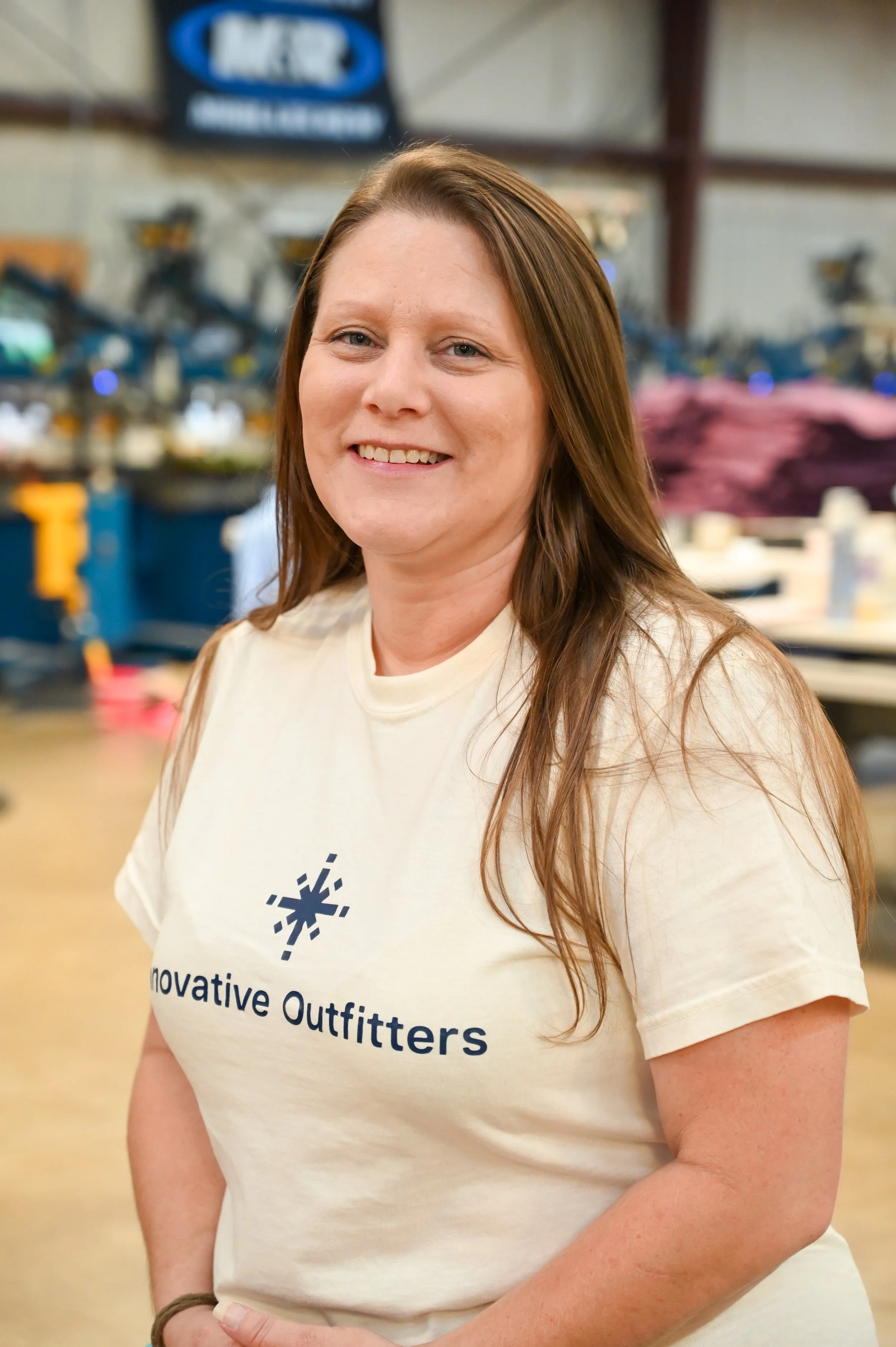 A smiling woman with long brown hair wearing a cream-colored T-shirt with the text 'Innovative Outfitters' and a snowflake logo, standing inside a warehouse or store with shelves and purple clothing in the background.