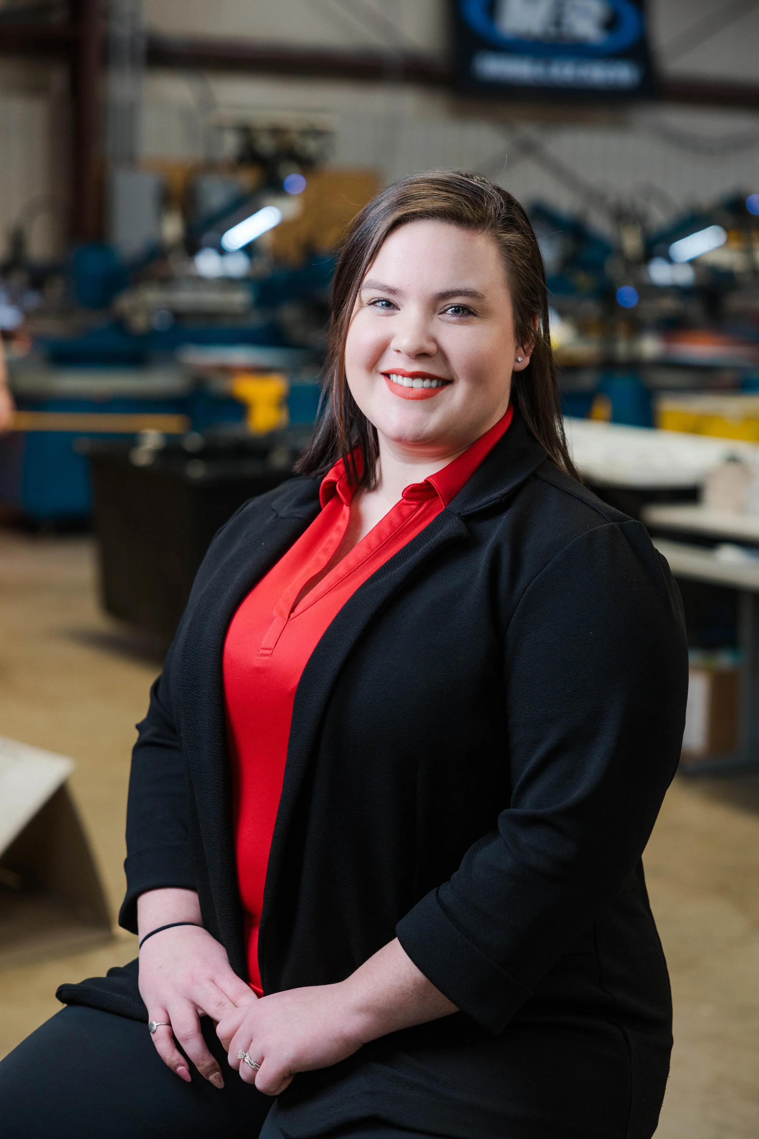 A woman smiling, seated in what appears to be a workshop or manufacturing setting, wearing a black blazer over a red shirt.