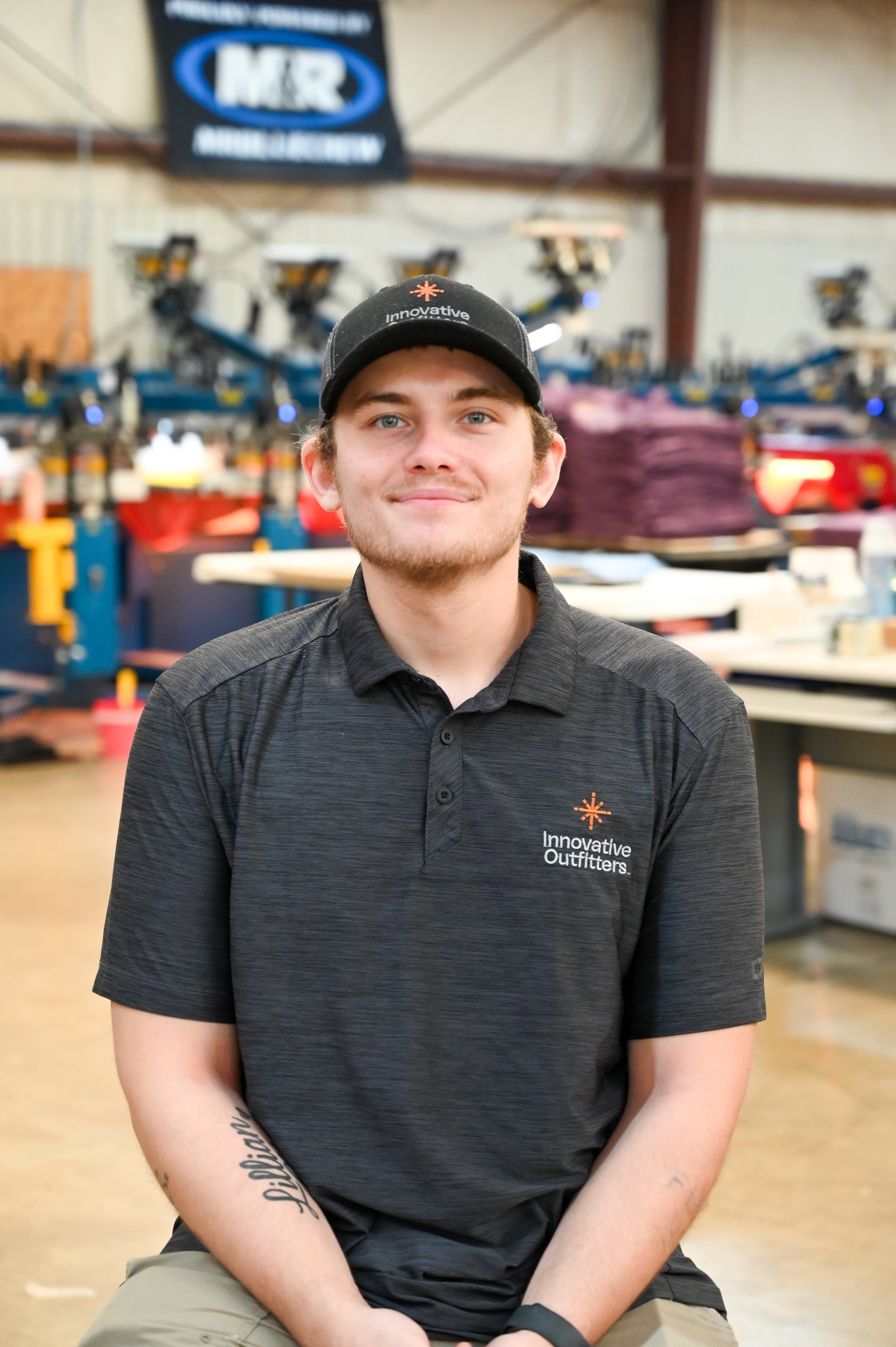 A young man with light brown hair and a beard, wearing a black baseball cap and a black polo shirt with 'Innovative Outfitters' logo, sitting in an industrial or warehouse environment with tools and equipment in the background.