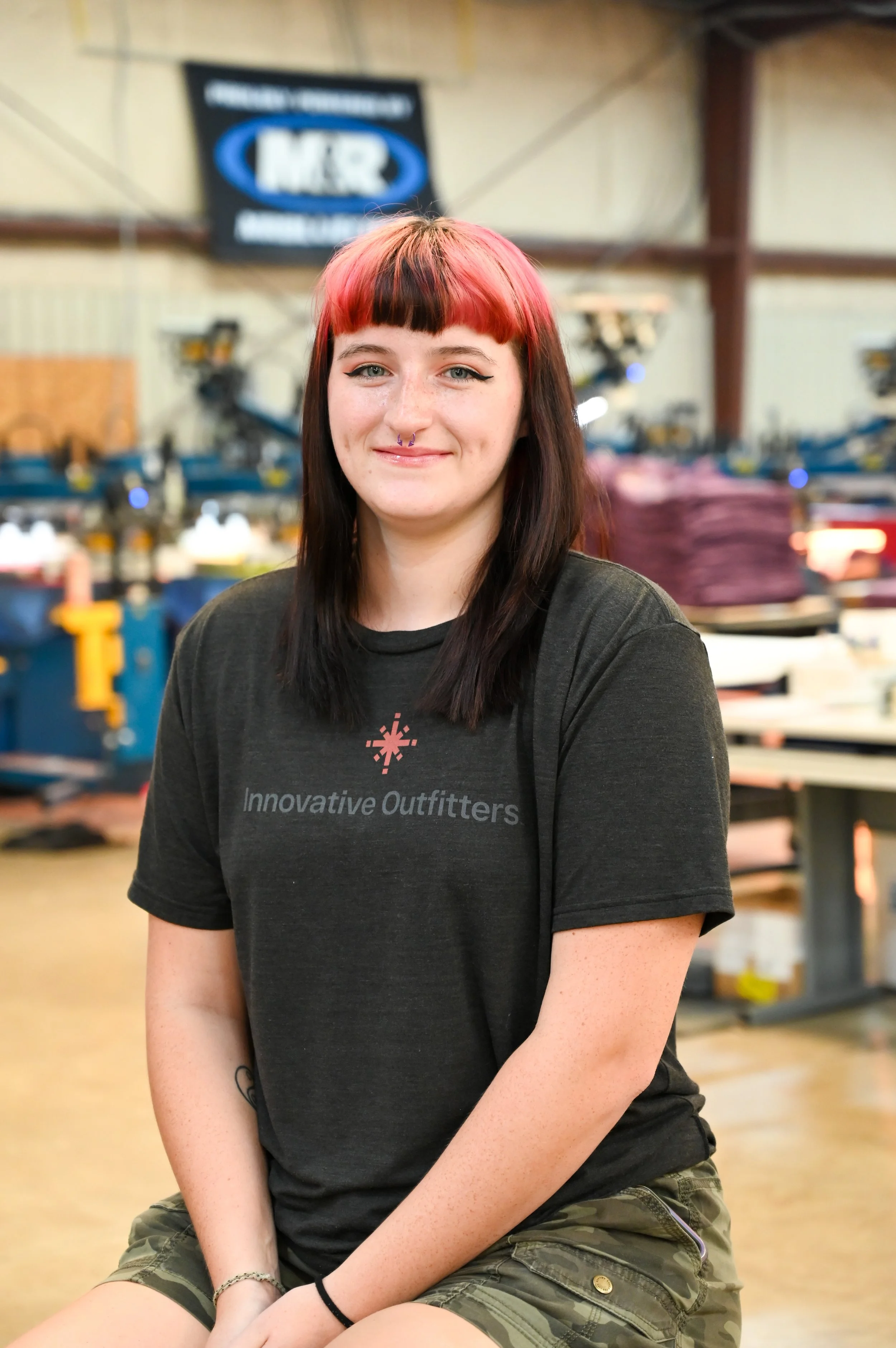 A young woman with red and black hair, wearing a black "Innovative Outfitters" t-shirt and camouflage shorts, sitting indoors with workshop equipment in the background.