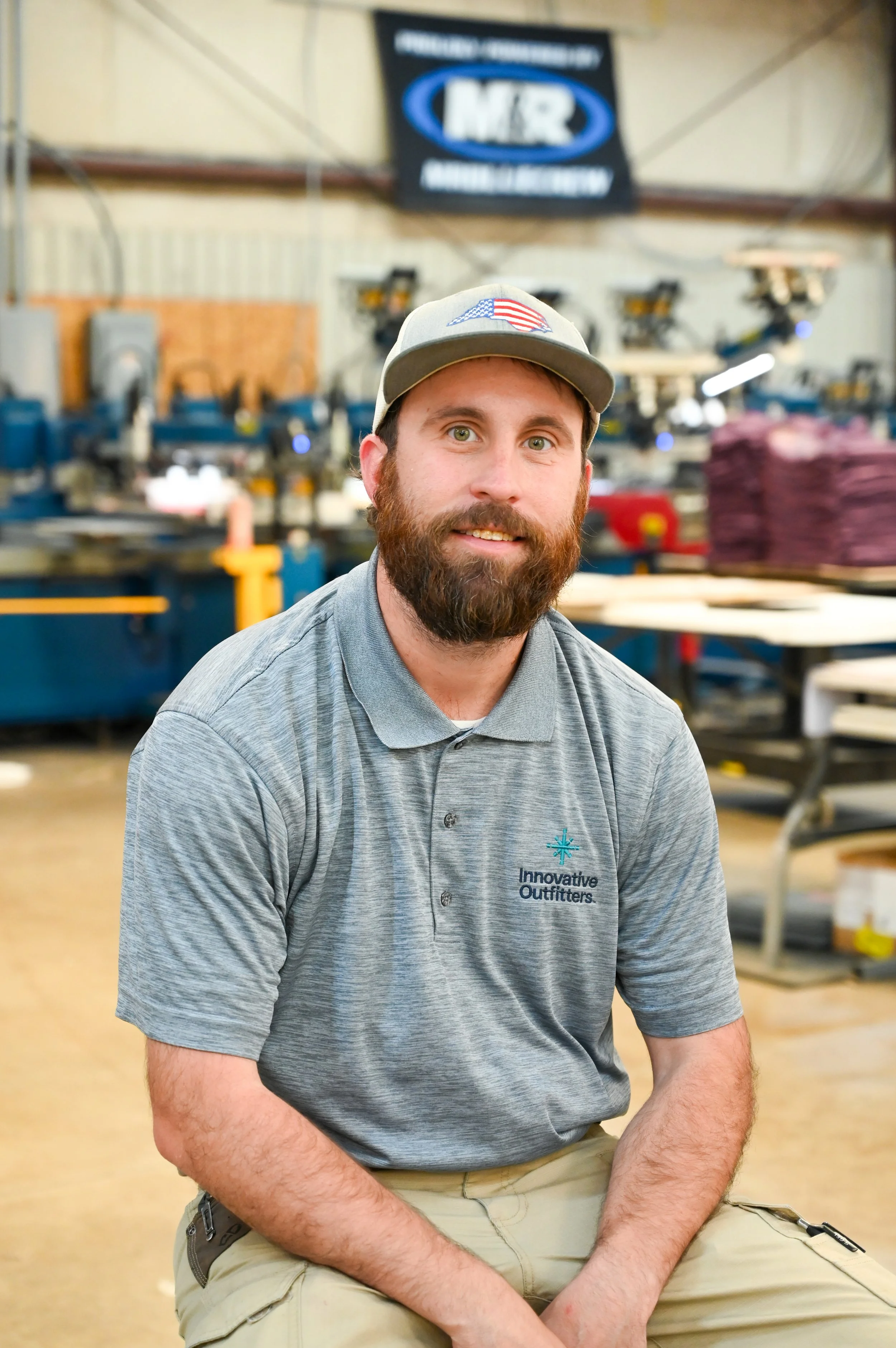 A man with a beard wearing a gray polo shirt with a logo that says "Innovative Outfitters" and a gray cap with an American flag patch, sitting in a woodworking or manufacturing shop with machinery and stacks of fabric or materials in the background.