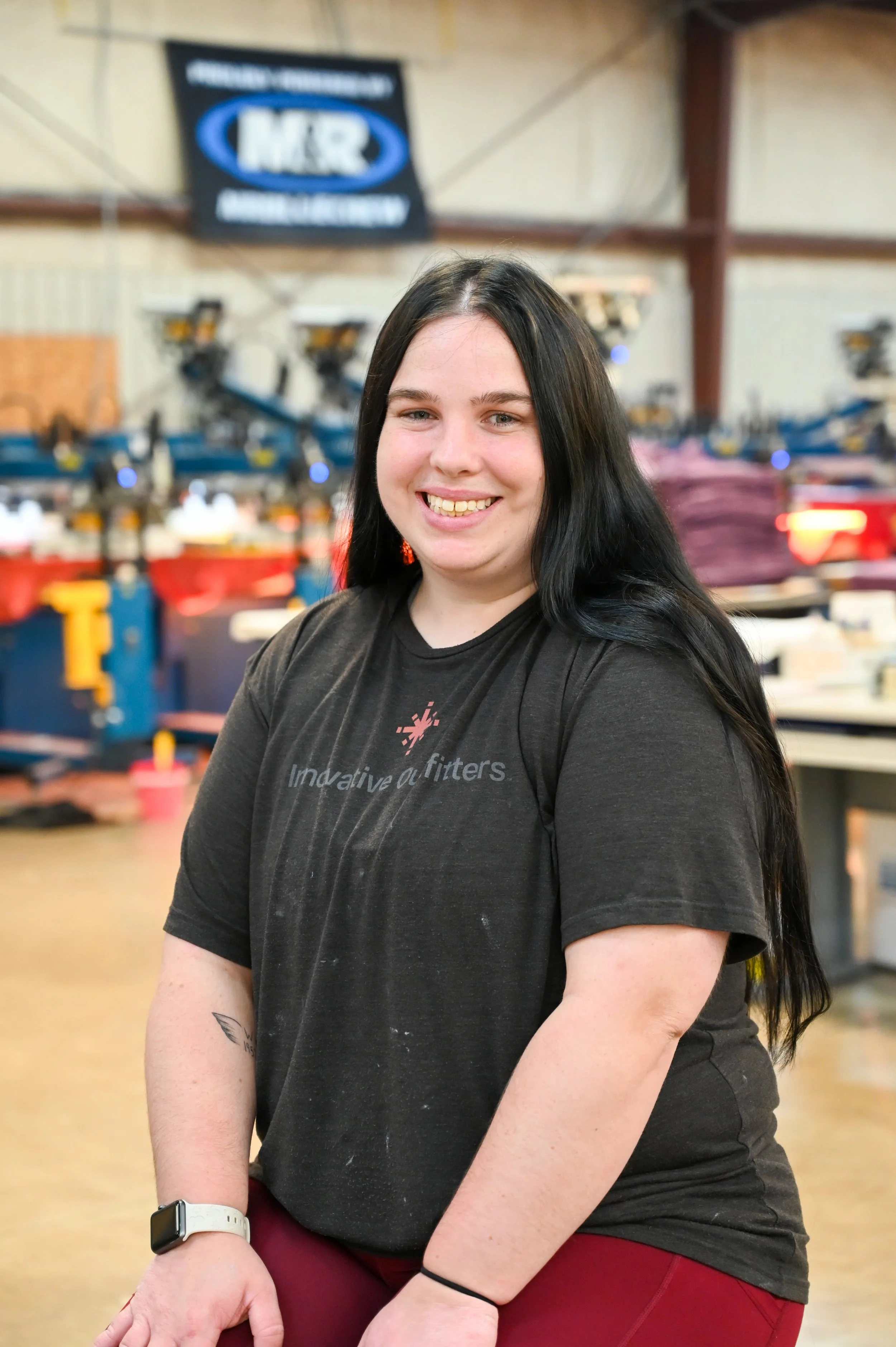 A woman with long black hair smiling and standing in a workshop or factory, with machinery and equipment in the background.