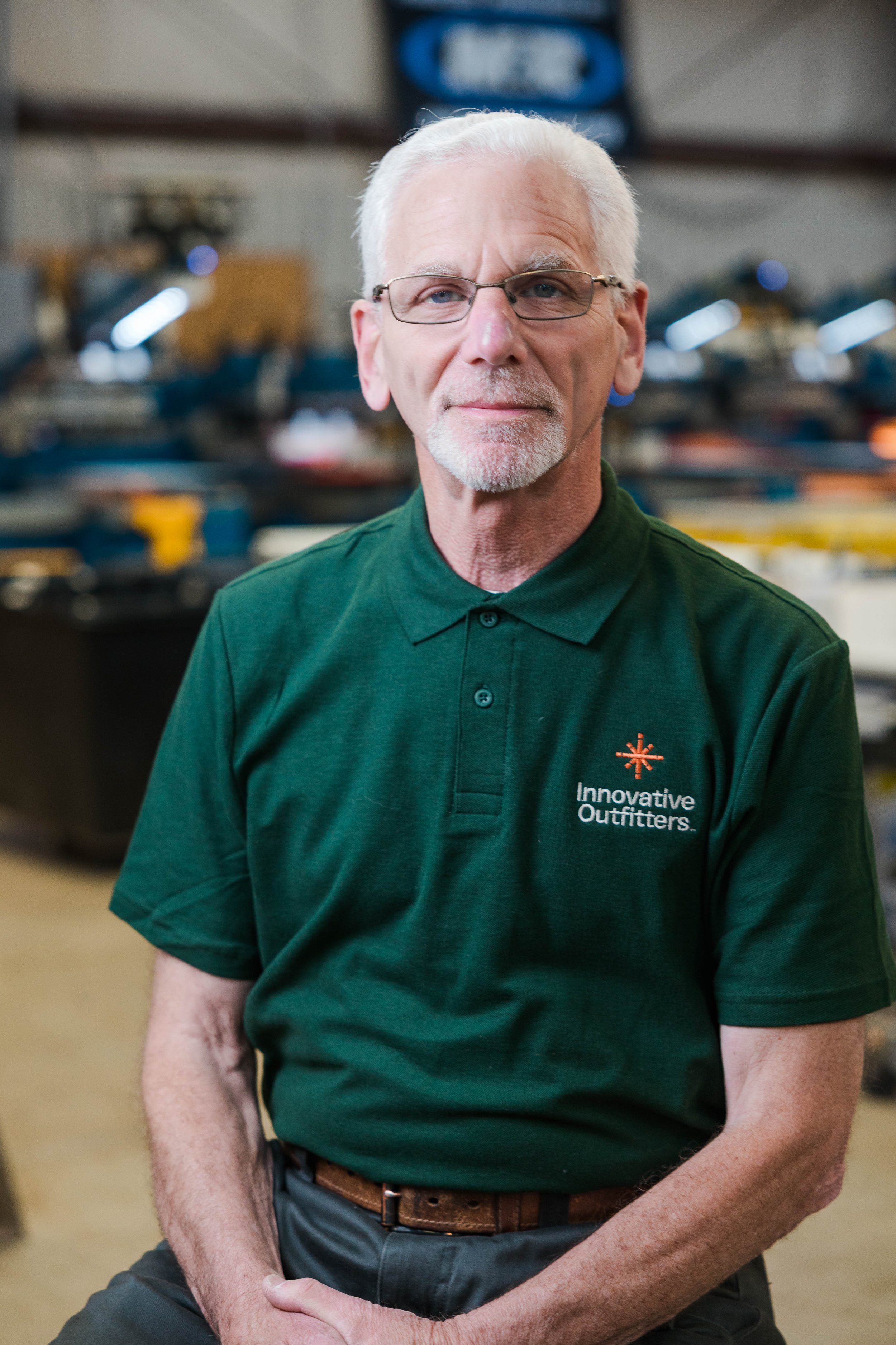 An older man with white hair, glasses, and a beard wearing a green polo shirt with the logo 'Innovative Outfitters' in an industrial or workshop setting.