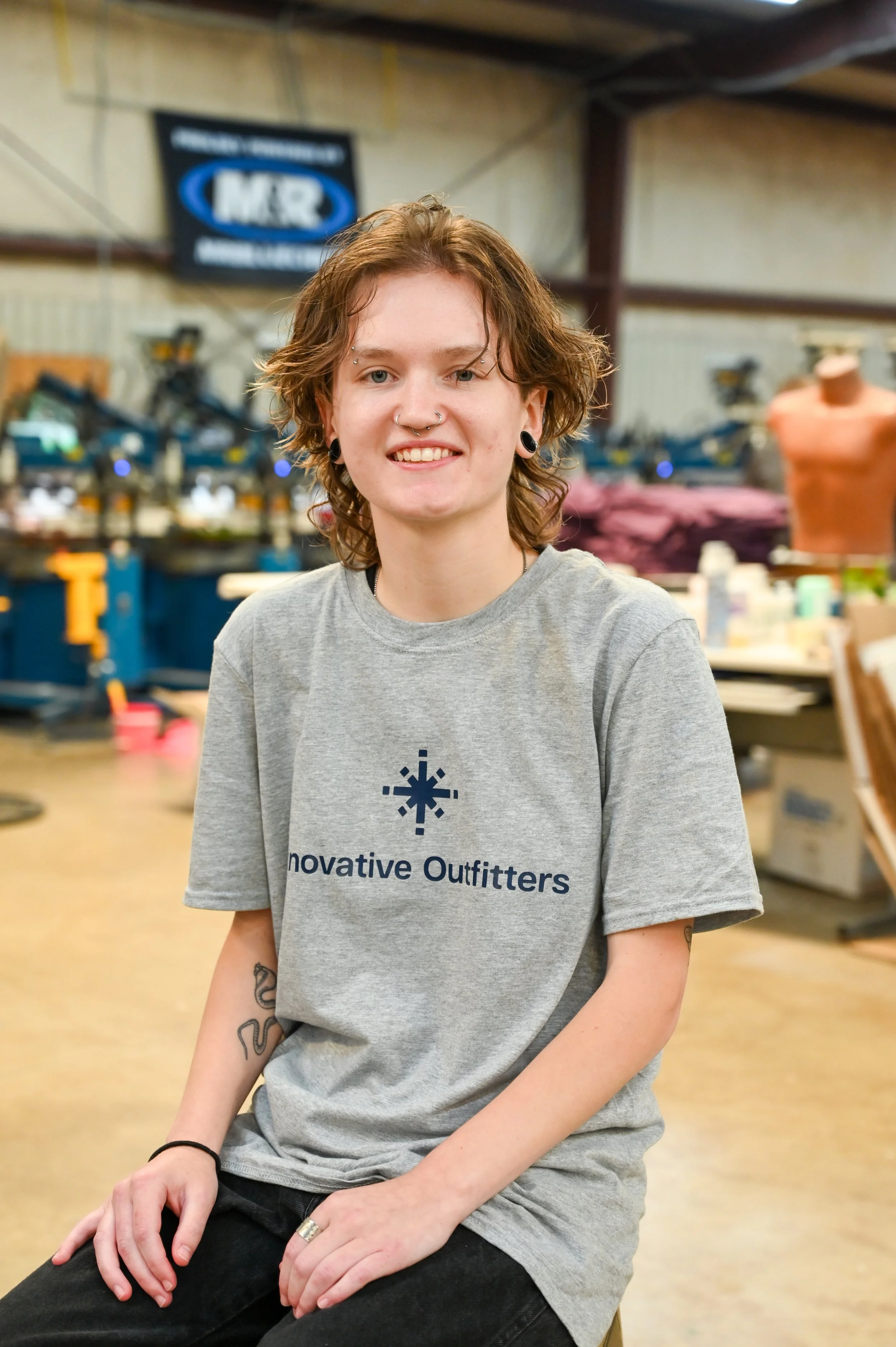 A young person with short, curly hair, multiple piercings, and tattoos on their arm, wearing a gray t-shirt with the words 'Innovative Outfitters' on it, sitting in an industrial workspace with machinery and a mannequin in the background.