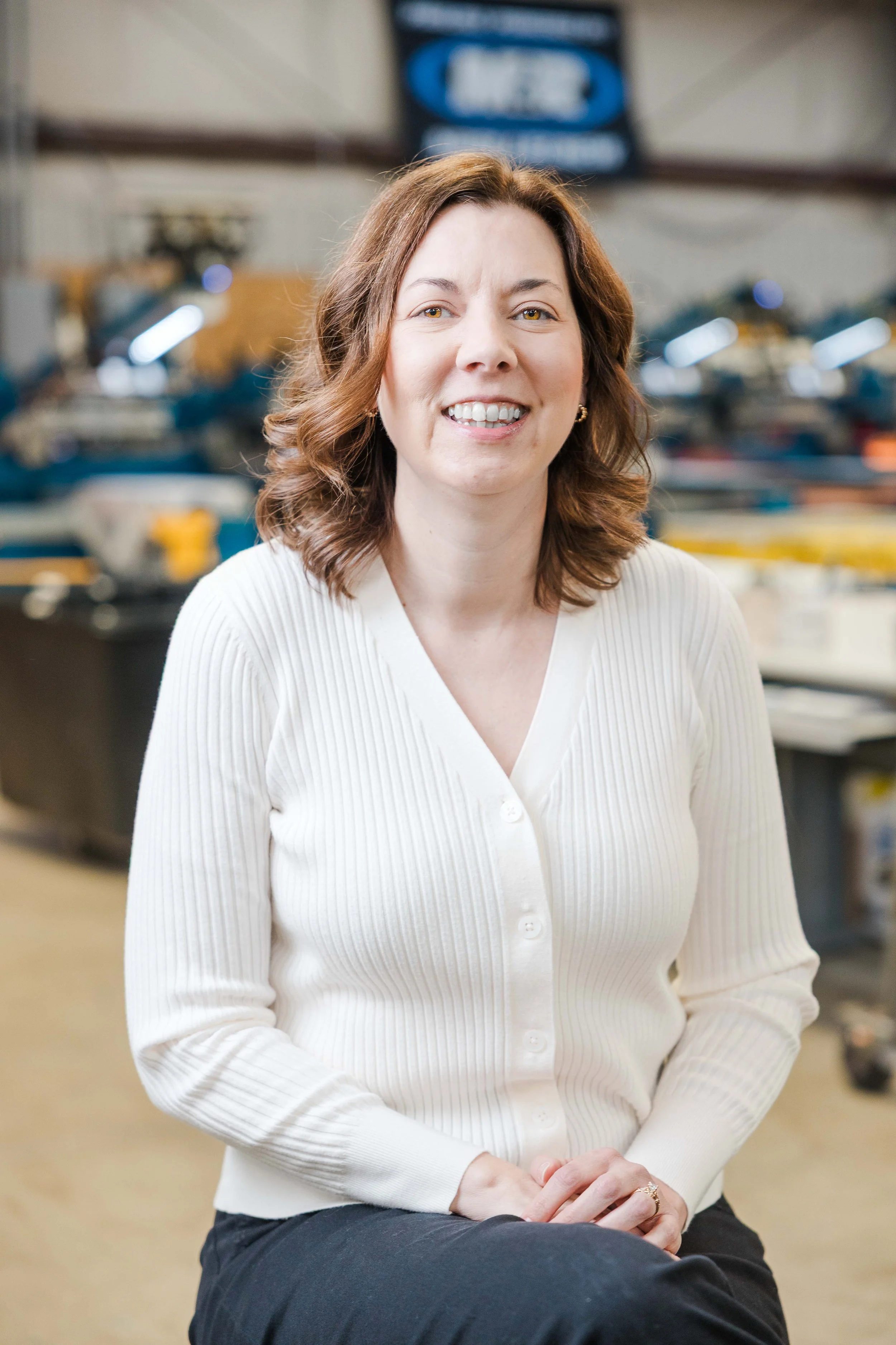 A woman with shoulder-length brown hair, wearing a white ribbed cardigan and black pants, sitting in an industrial setting with machinery and equipment in the background, smiling at the camera.