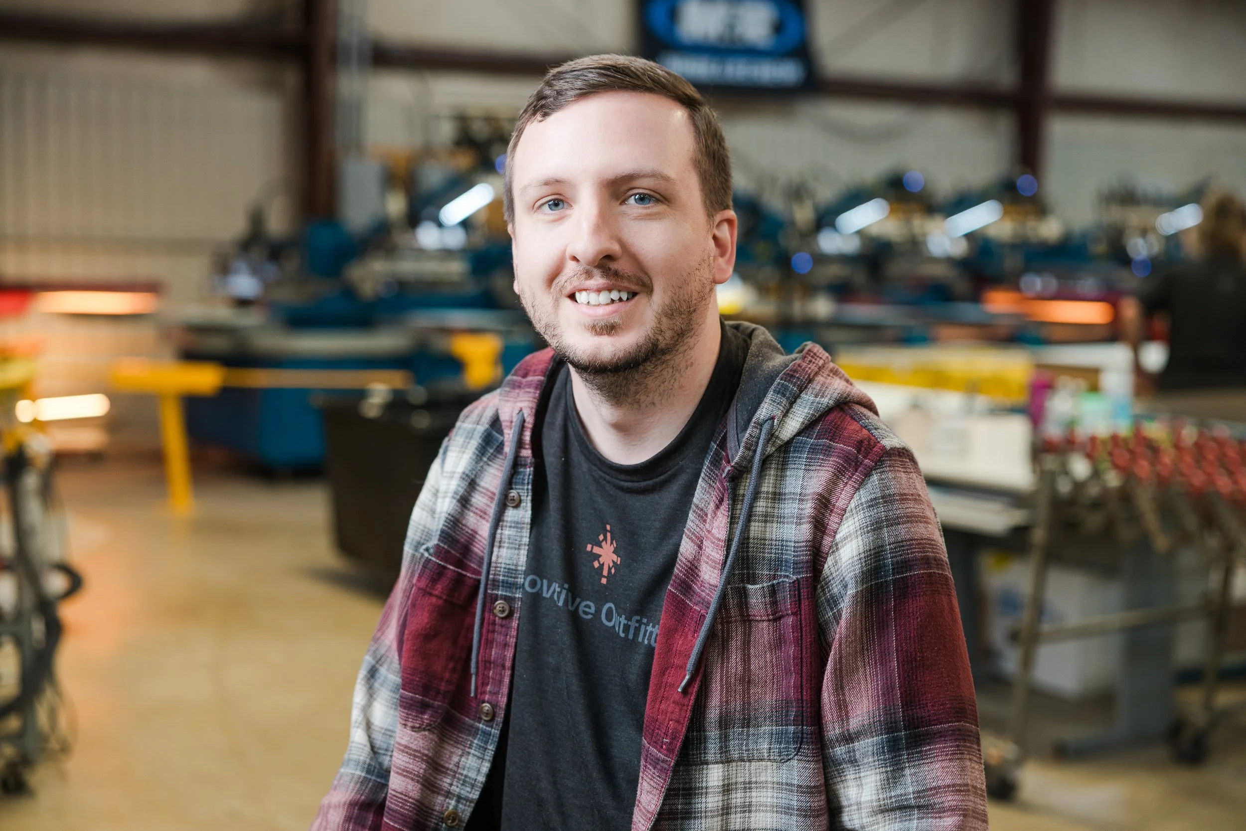 A young man with short brown hair and a beard, smiling, wearing a black t-shirt and a plaid hoodie, standing inside a kayak shop or warehouse with rows of kayaks and equipment in the background.
