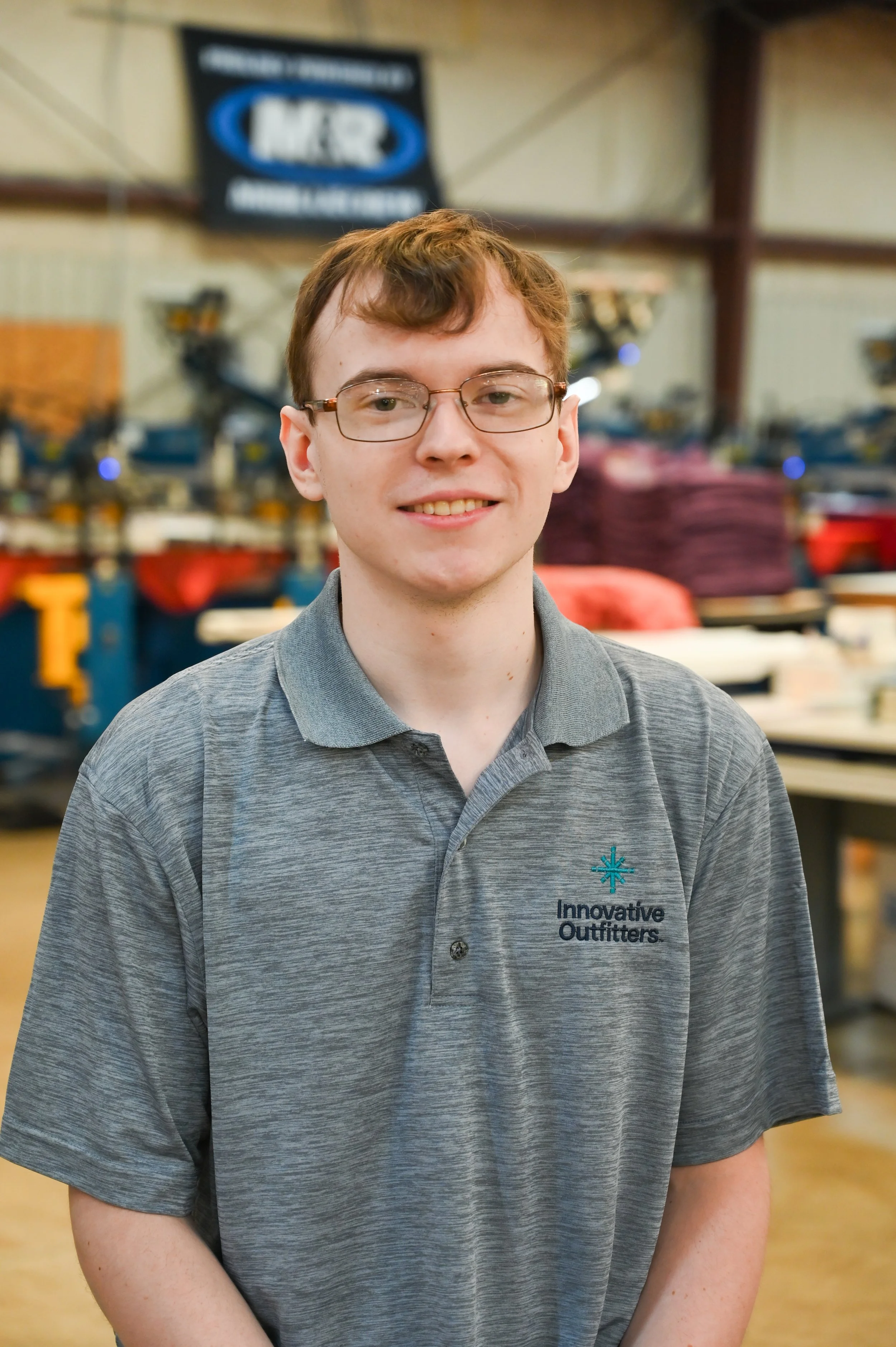 A young man wearing glasses and a gray polo shirt with 'Innovative Outfitters' logo, smiling indoors in a workshop or factory environment.