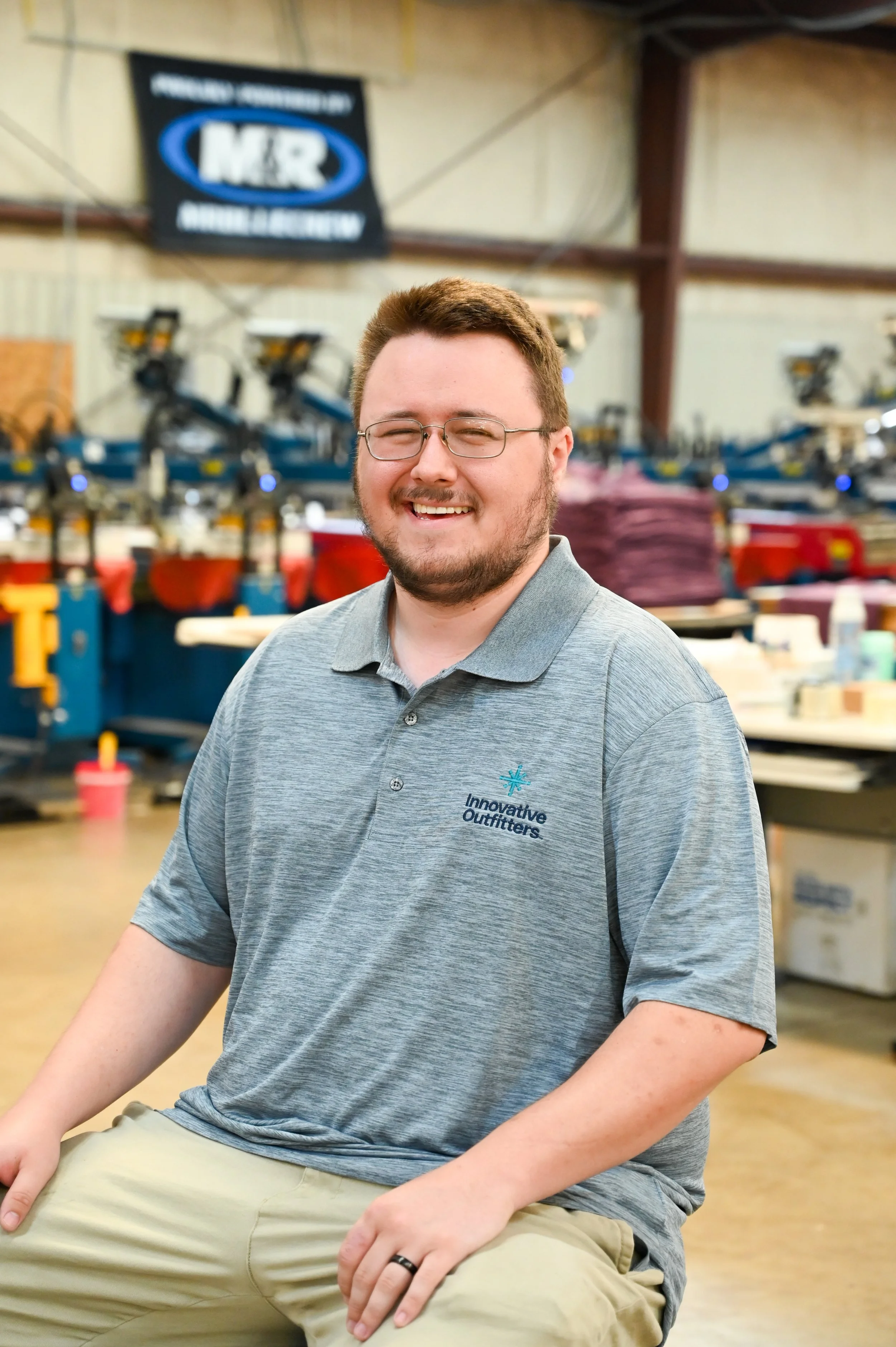 A smiling man with glasses and a beard wearing a gray polo shirt with 'Innovative Outfitters' logo, sitting indoors with a blurred industrial or workshop background.