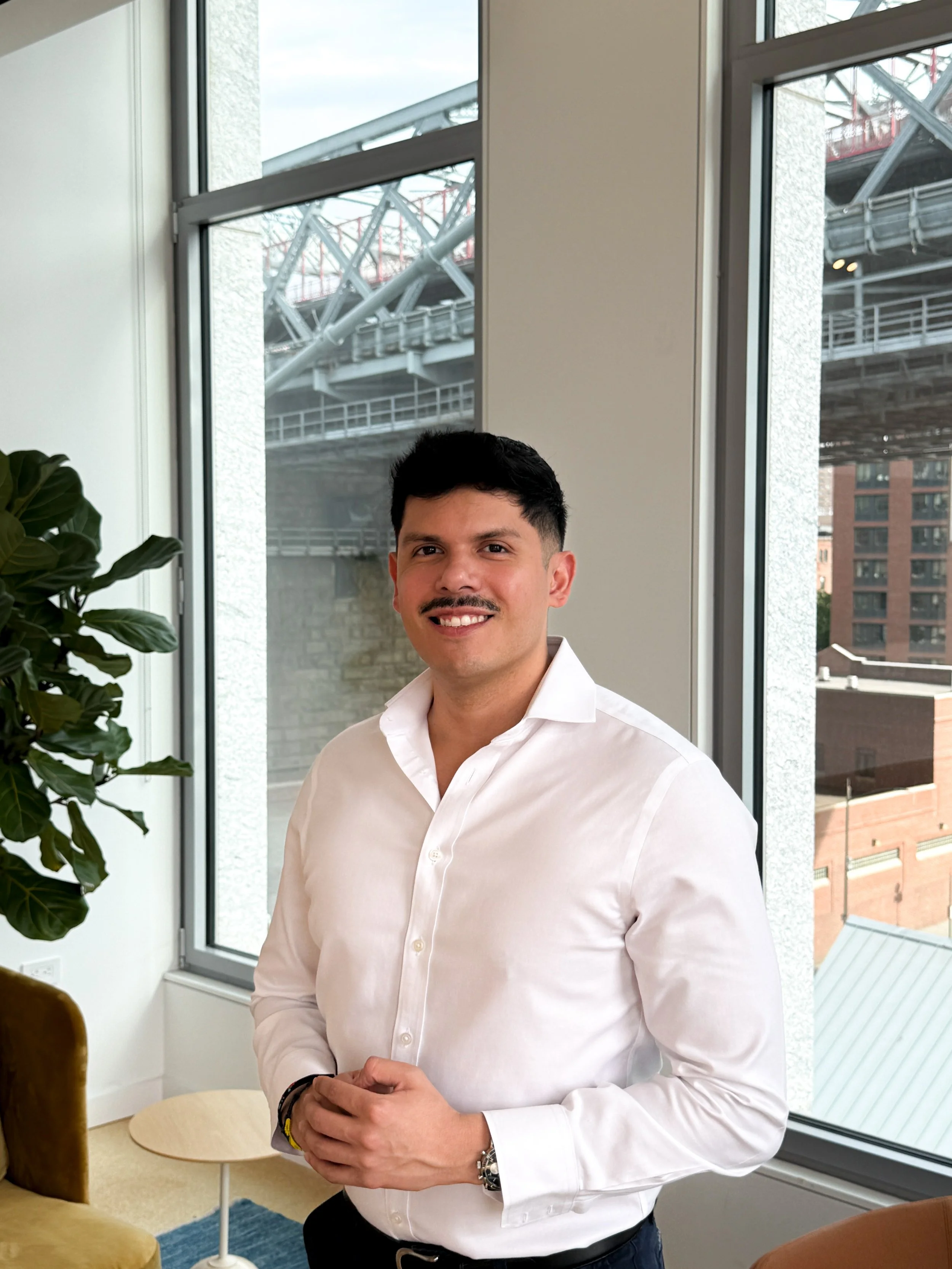 A man with dark hair and a mustache wearing a white dress shirt, standing indoors next to large windows overlooking a cityscape with buildings and an elevated highway.