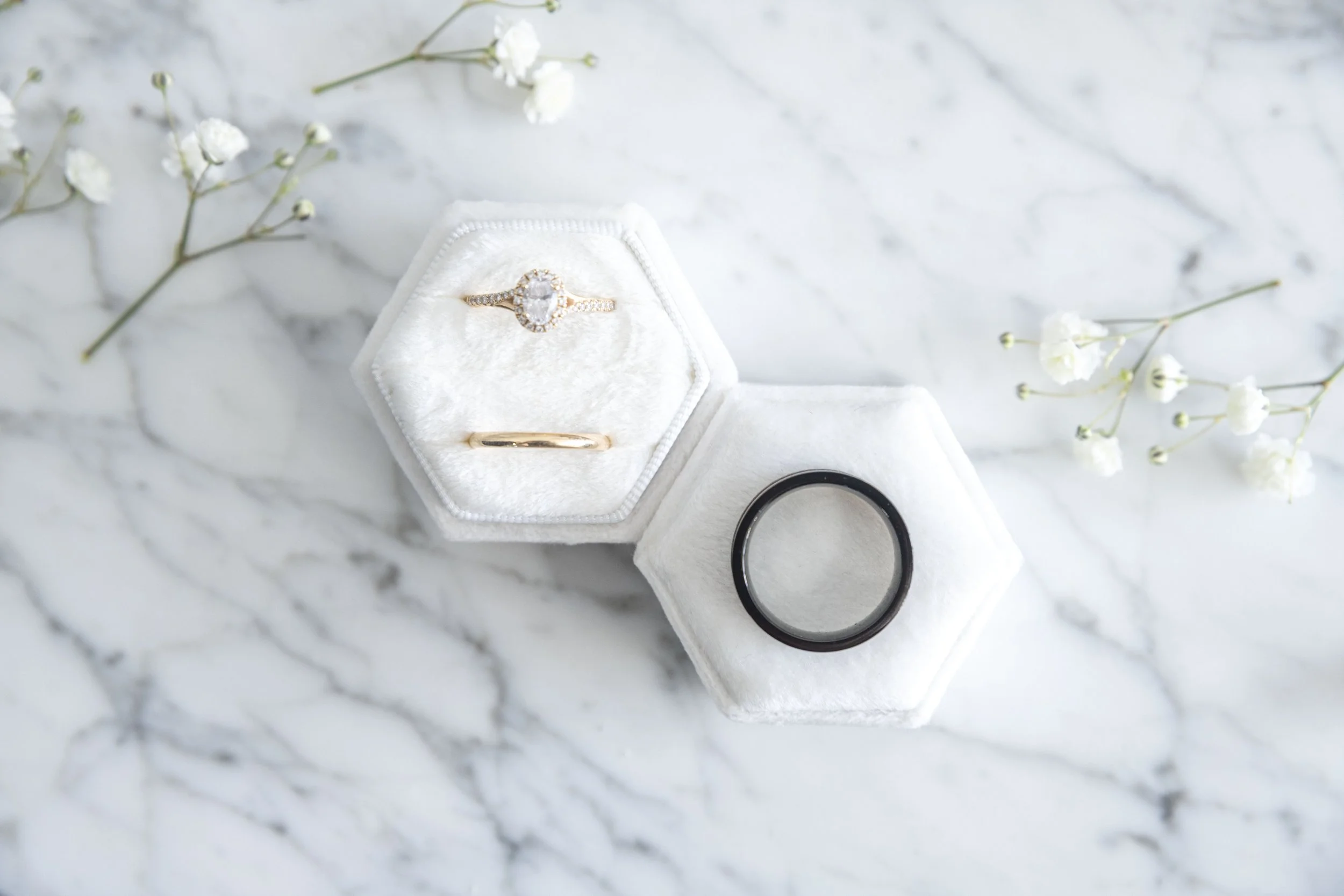 A white jewelry ring box containing a diamond engagement ring and a wedding band, placed on a white marble surface with small white flowers nearby.