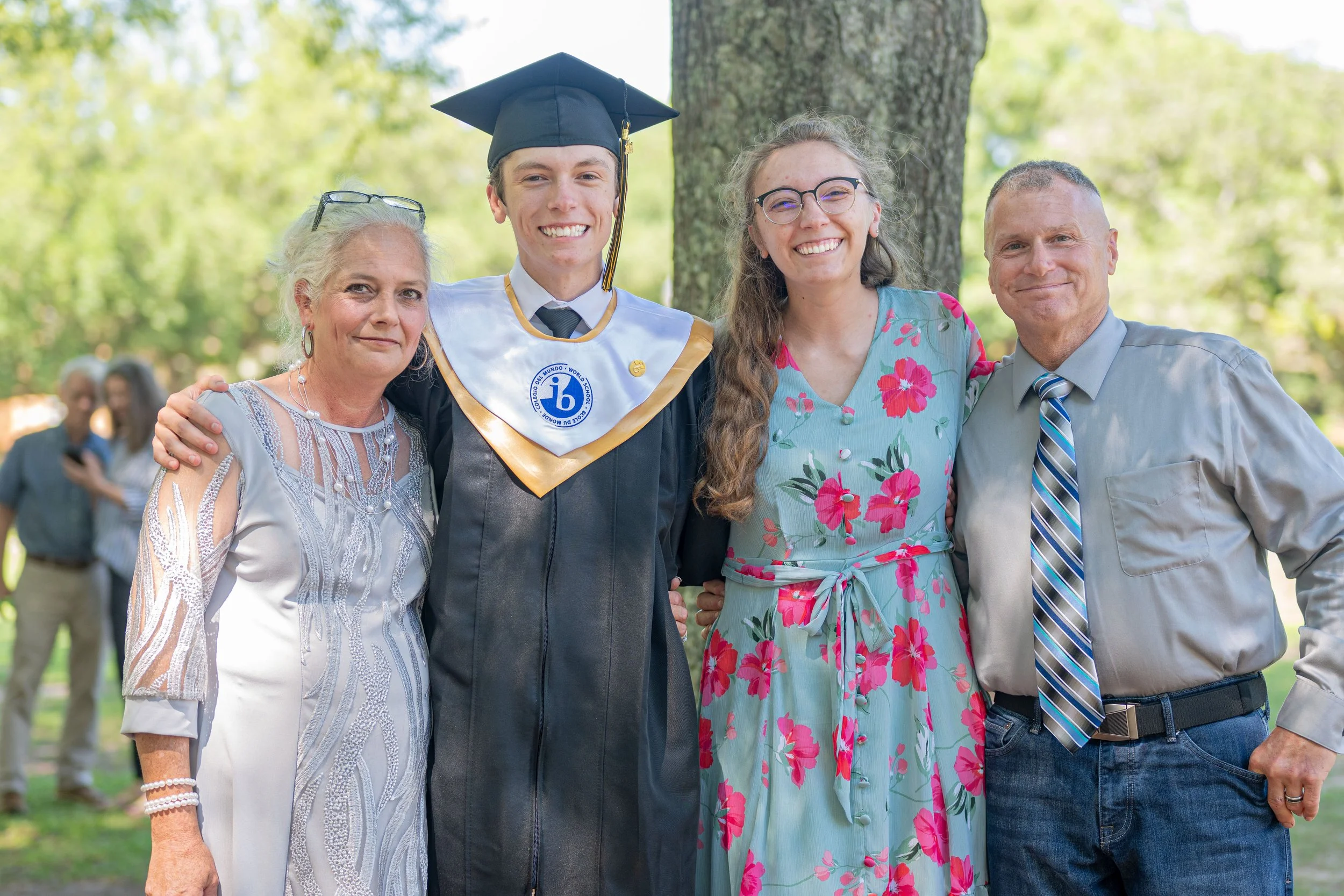 A young man in a graduation cap and gown with a white and blue collar, standing with three women and a man outdoors on a sunny day. The women are dressed in light-colored dresses, and the man in a gray shirt with a striped tie. The background shows t