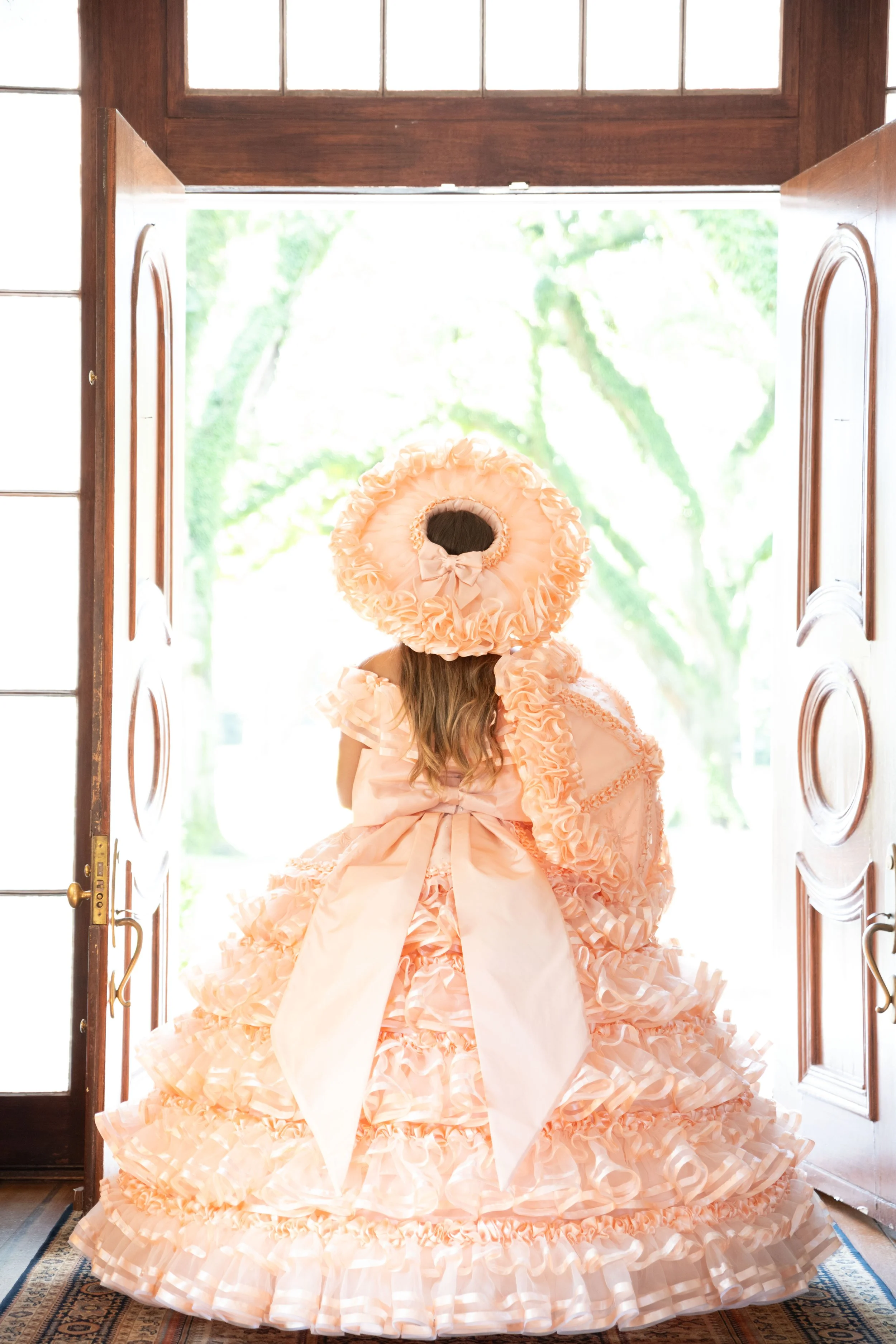 Person in peach ruffled dress and matching bonnet standing in open doorway.