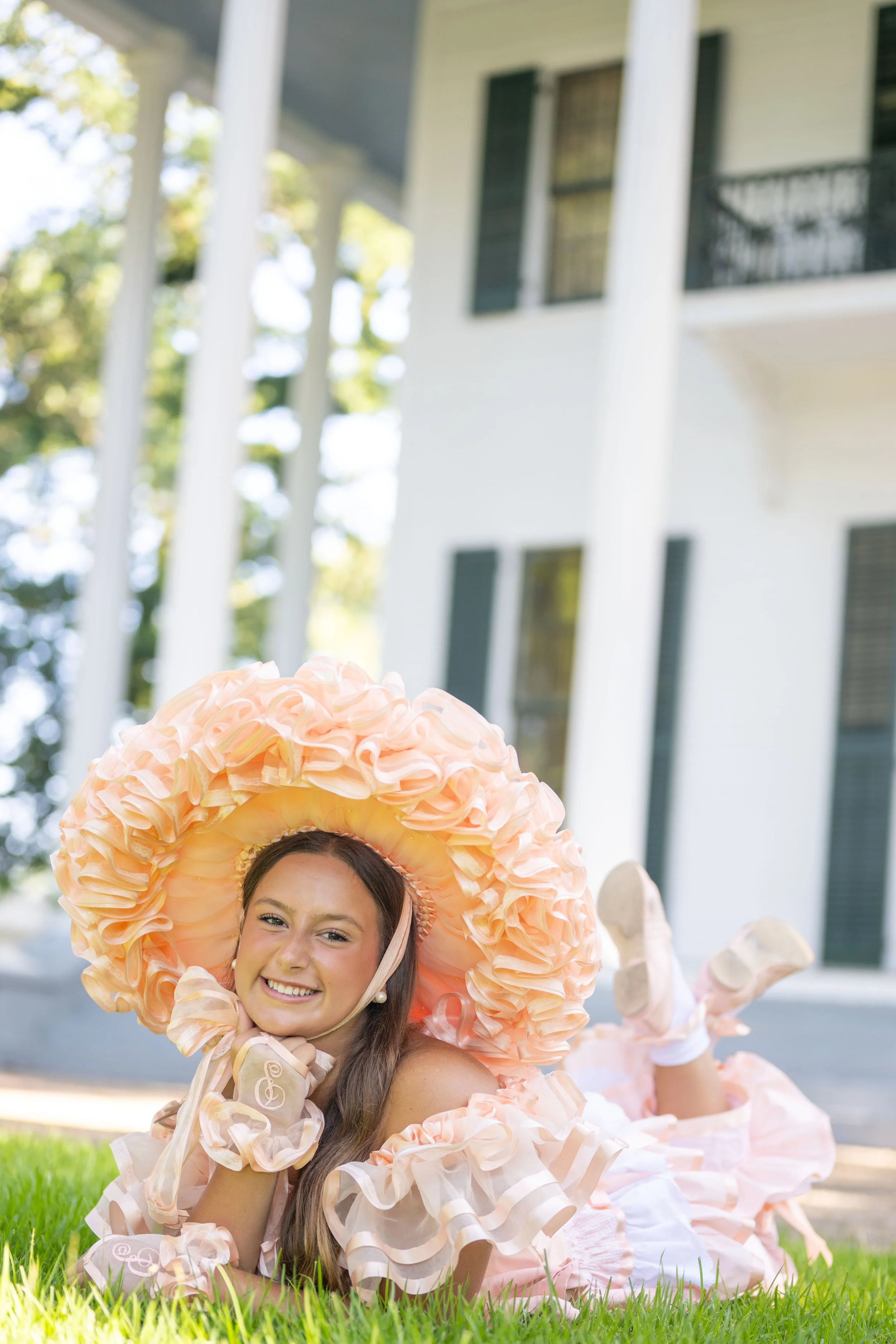 Young woman in an orange bonnet and frilly dress lying on the grass in front of a large white house with columns.
