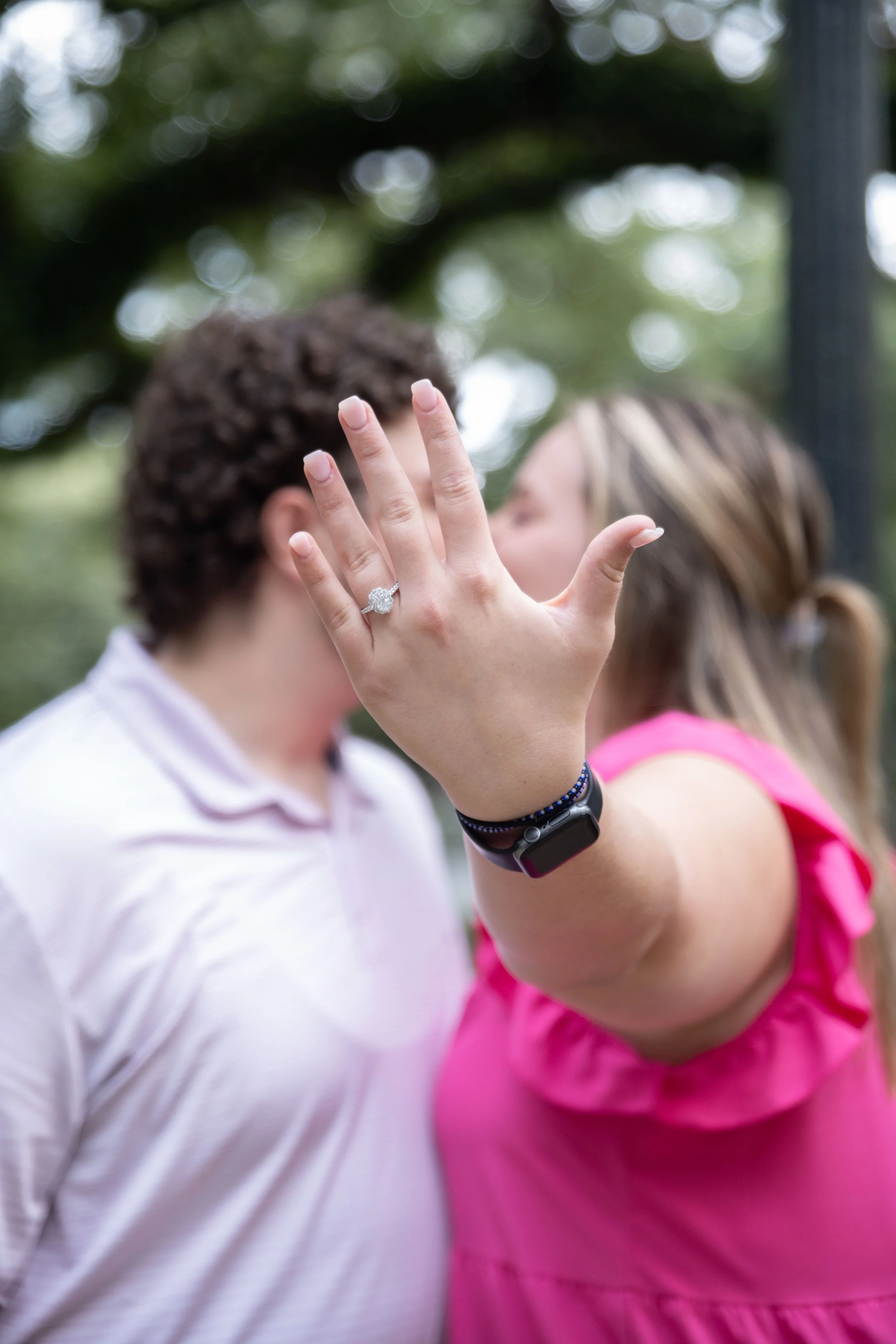 Close-up of a woman's hand showing an engagement ring, with a couple blurred in the background.