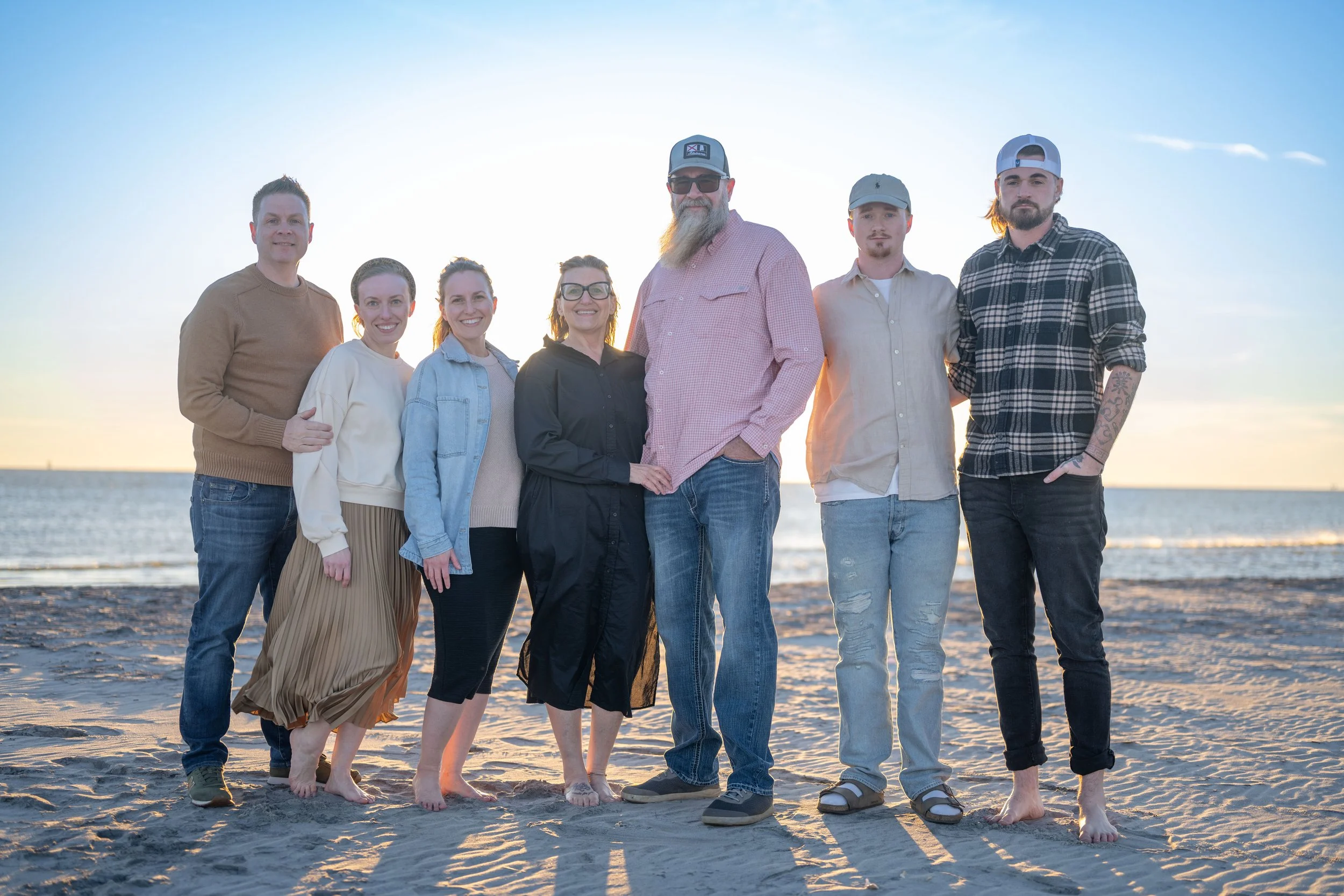 Group of seven people standing barefoot on a beach during sunset, with the ocean in the background.
