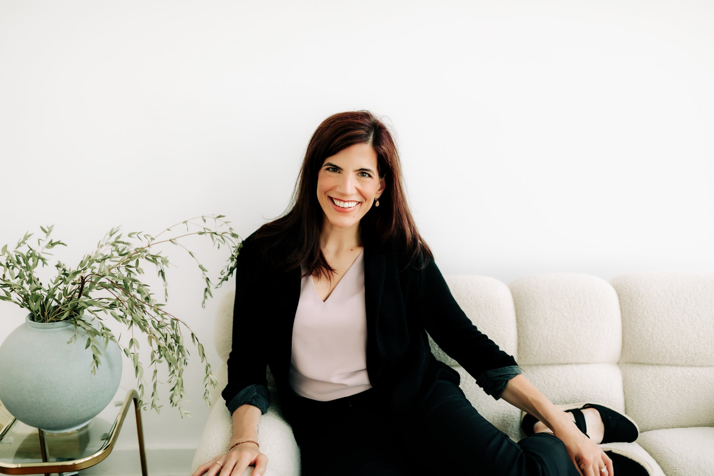 Smiling woman with dark hair sitting on a cream-colored sofa next to a green potted plant in a modern room.