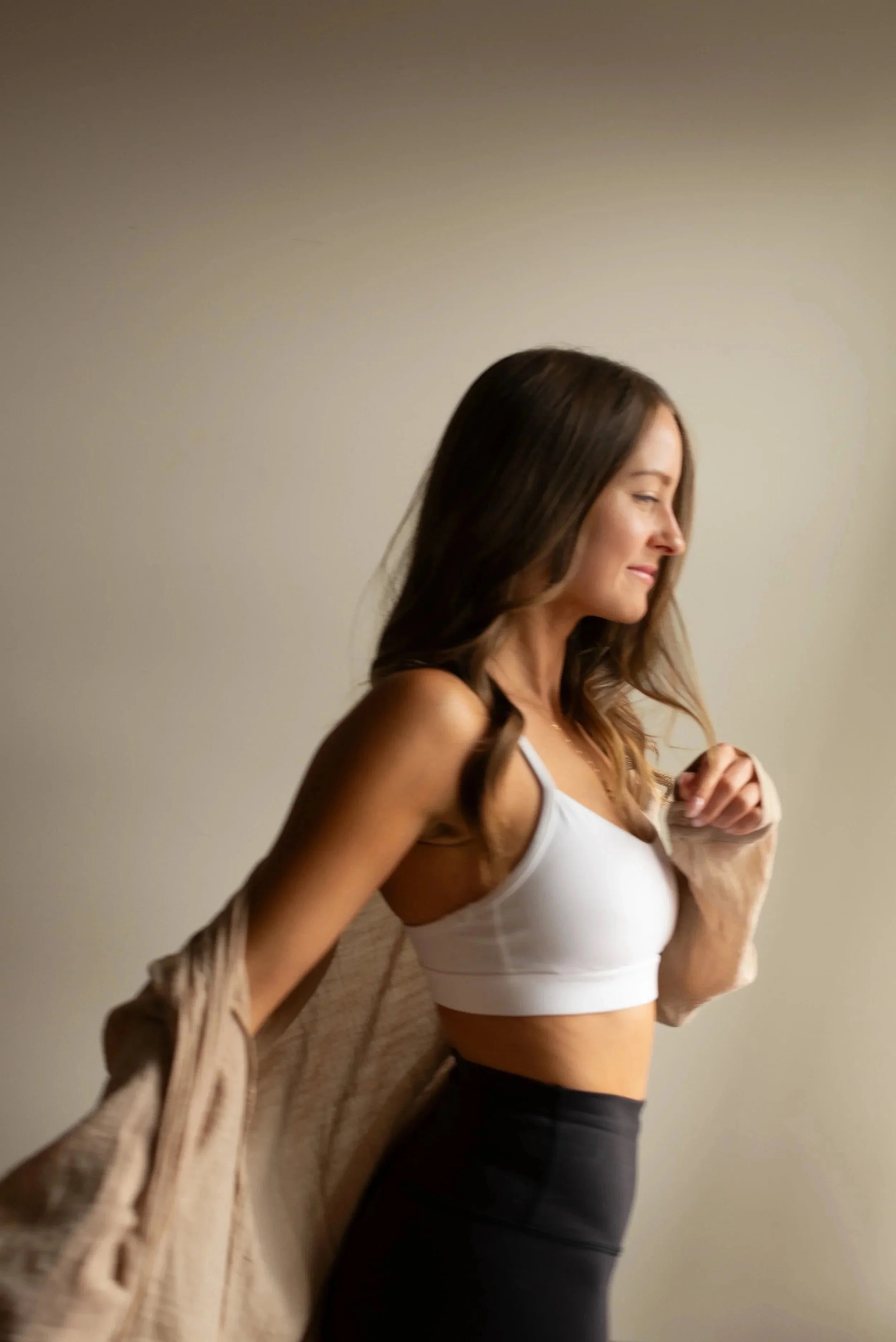 A woman with long, wavy brown hair wearing a white sports bra and black leggings, standing against a plain beige wall, holding a beige jacket over her shoulder and looking to the side with a slight smile.