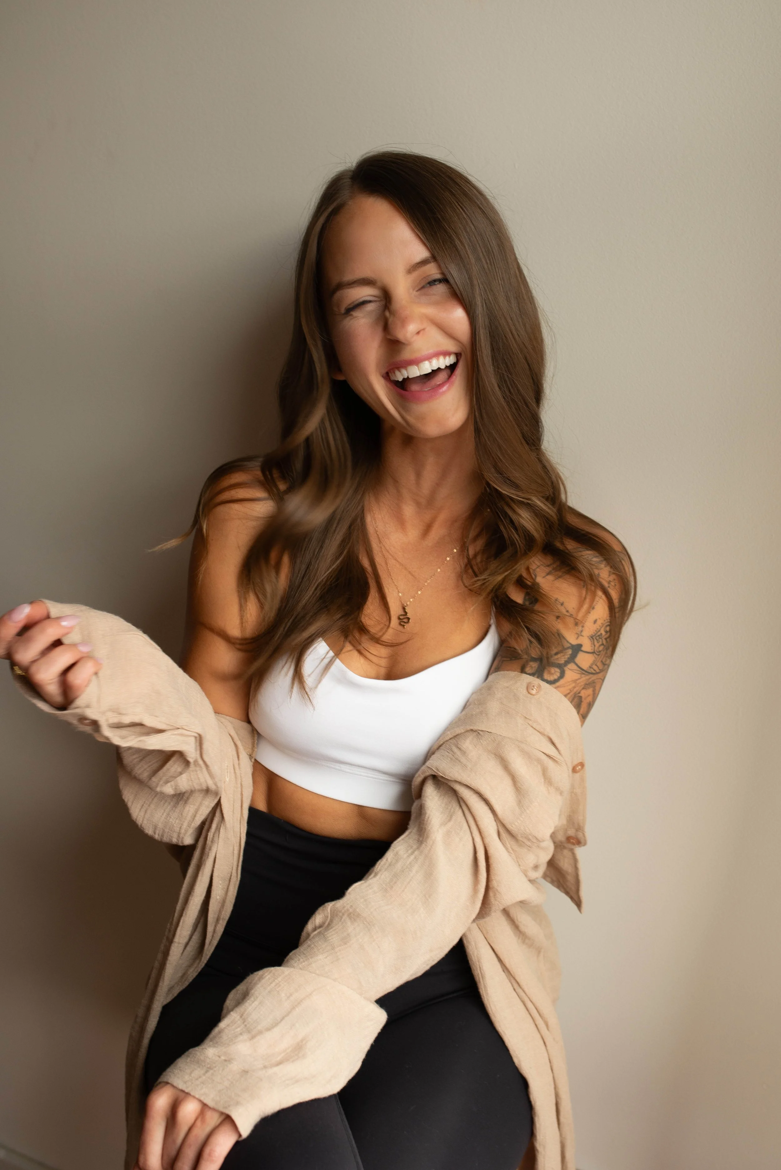 A woman with long, wavy brown hair, smiling and laughing, wearing a white crop top, black pants, and a beige jacket, sitting against a plain beige wall.