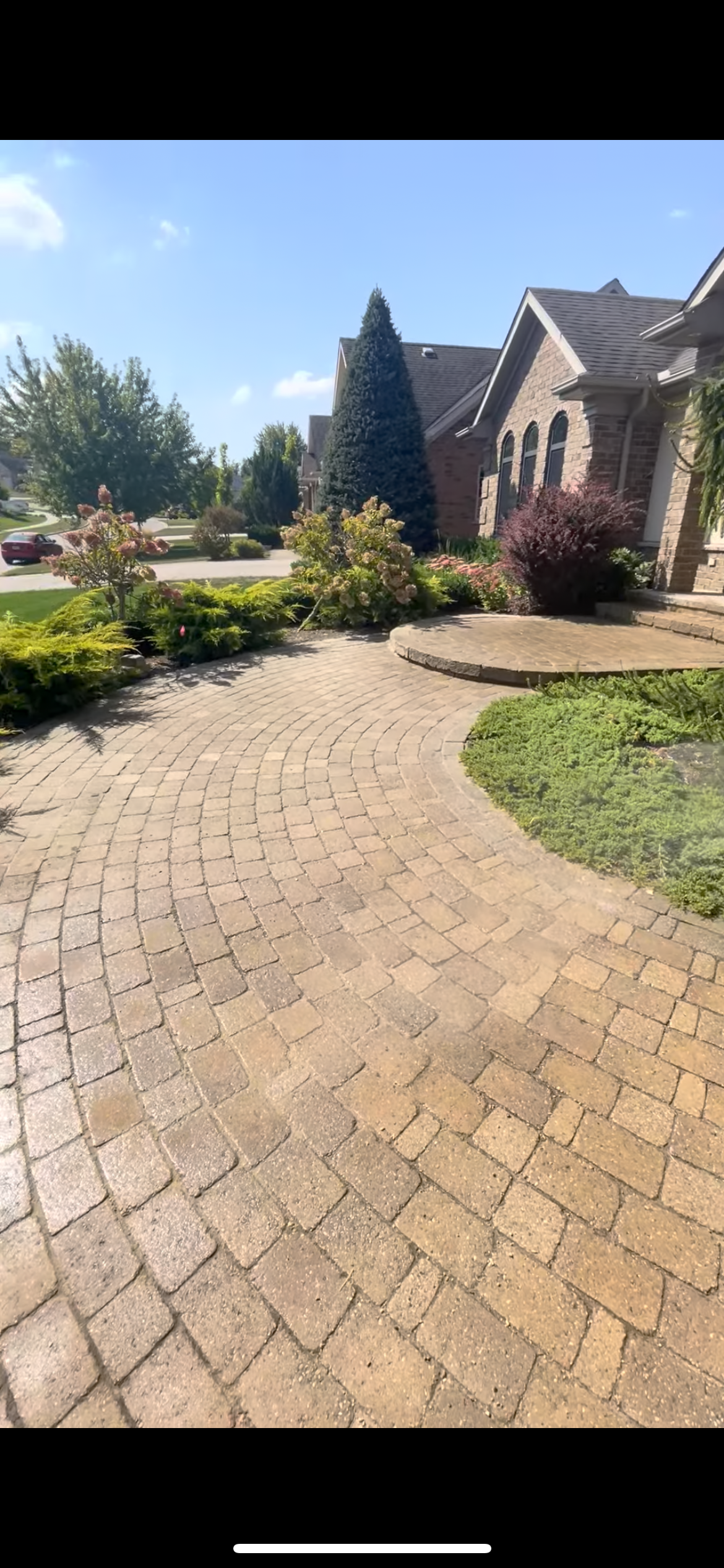 A curved brick walkway leading to the front steps of a suburban house with well-maintained garden landscaping and a clear blue sky.