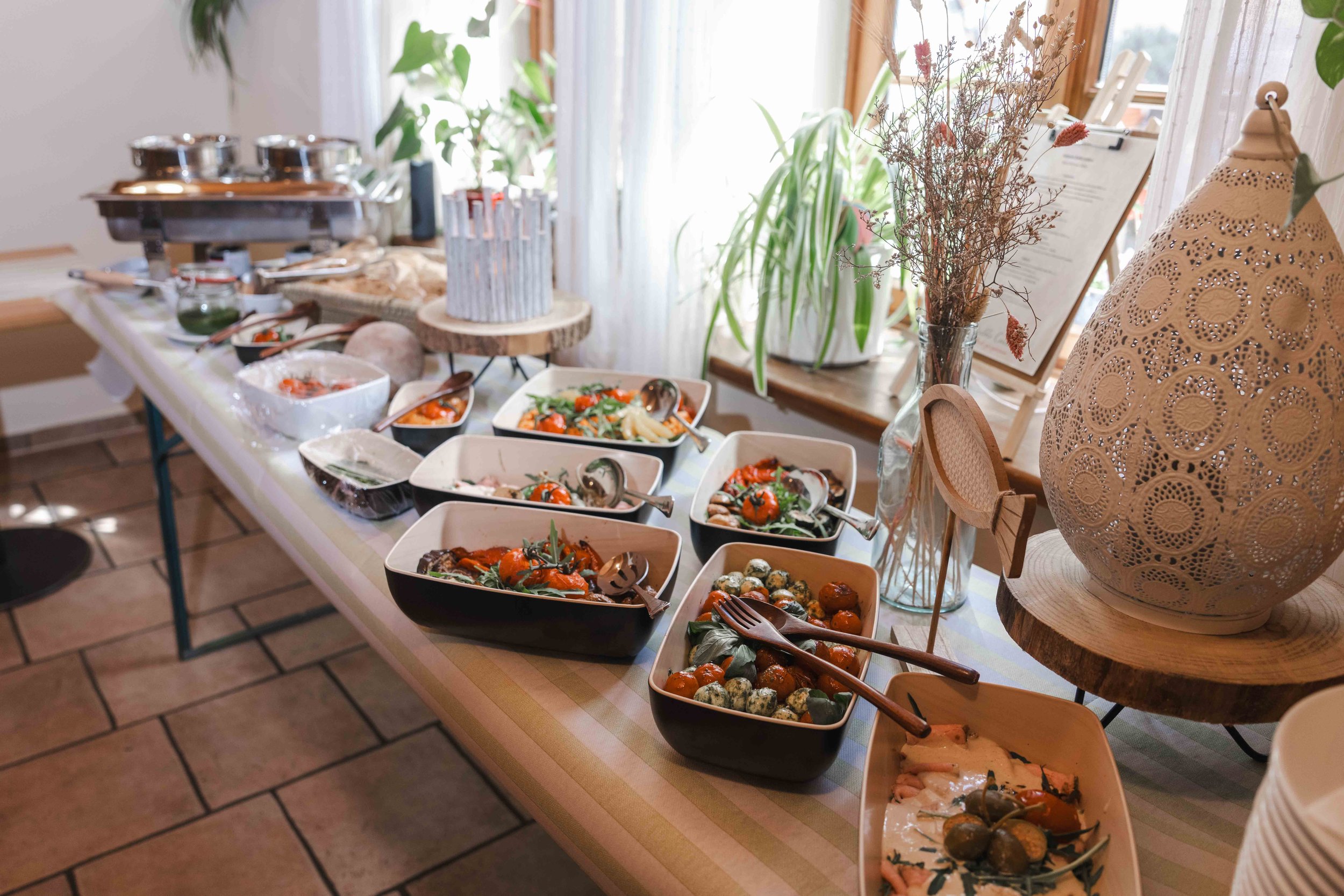 Buffet mit verschiedenen Gerichten in Schalen, Beleuchtung, Tischdekoration und grünen Pflanzen im Hintergrund.