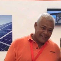 A smiling man with short gray hair wearing an orange shirt standing indoors with a solar panel display in the background.