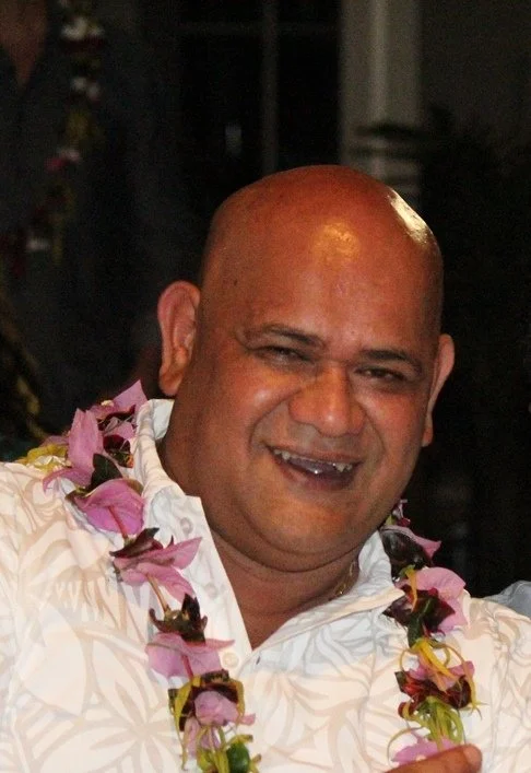 A smiling man with a shaved head wearing a white floral shirt and a pink lei around his neck.