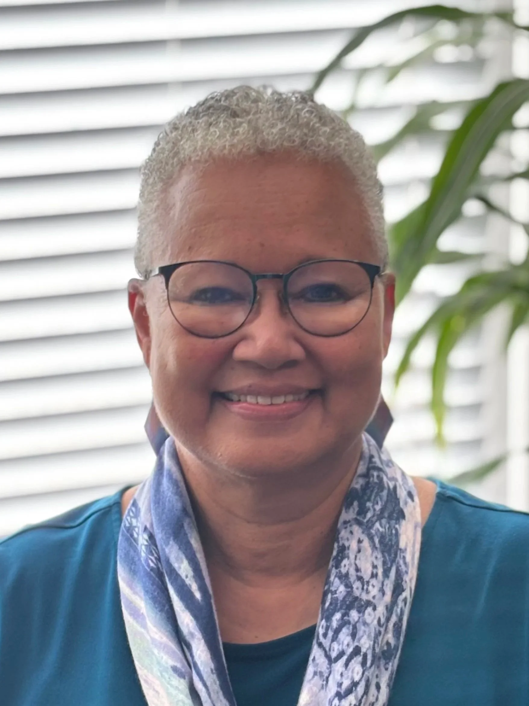 A smiling person with short, curly gray hair wearing glasses and a patterned scarf around their neck, standing in front of a background with horizontal blinds and green plants.