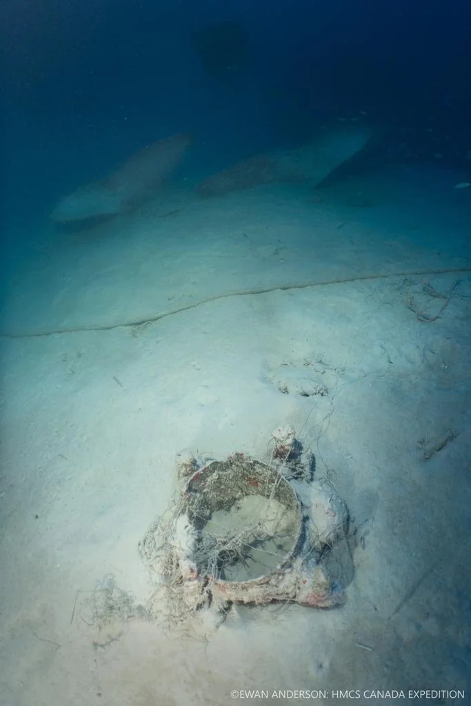 A broken deck fitting (foreground), tangled in fishing line, within the starboard-side debris field near the pair of spare propellers (background).