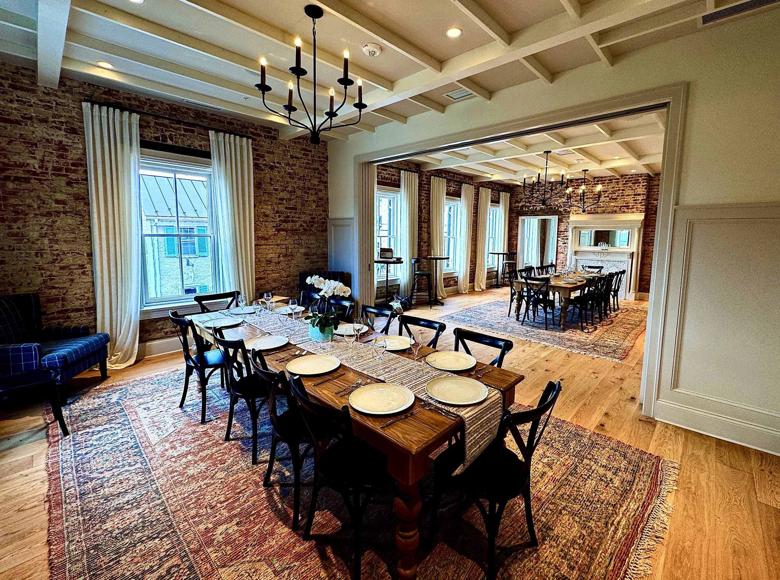 Elegant dining room with exposed brick walls, large windows with white curtains, hardwood floors, and multiple wooden tables set with white plates, wine glasses, and silverware, accented by black chairs and vintage-style chandeliers.
