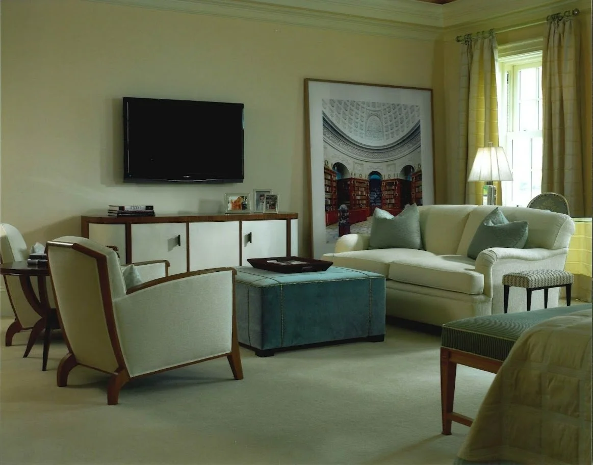Modern living room with a beige sofa, accent chairs, and a blue ottoman. A flat-screen TV is mounted on the wall above a sideboard with books and framed photos. Large framed artwork and a lamp on a table near a window with curtains complete the decor