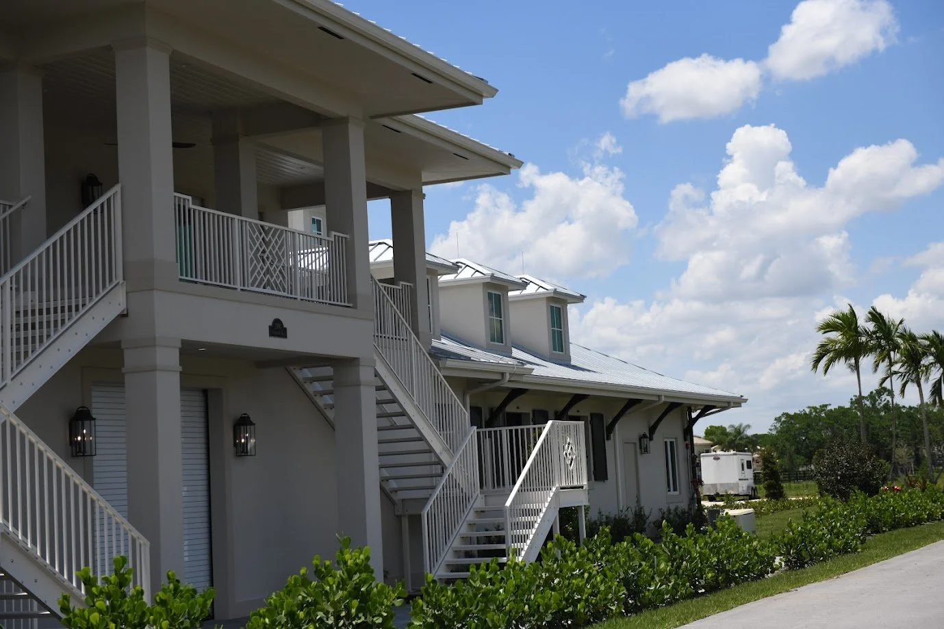 Modern white horse stable with external staircases and metal roof, surrounded by greenery, on a sunny day.