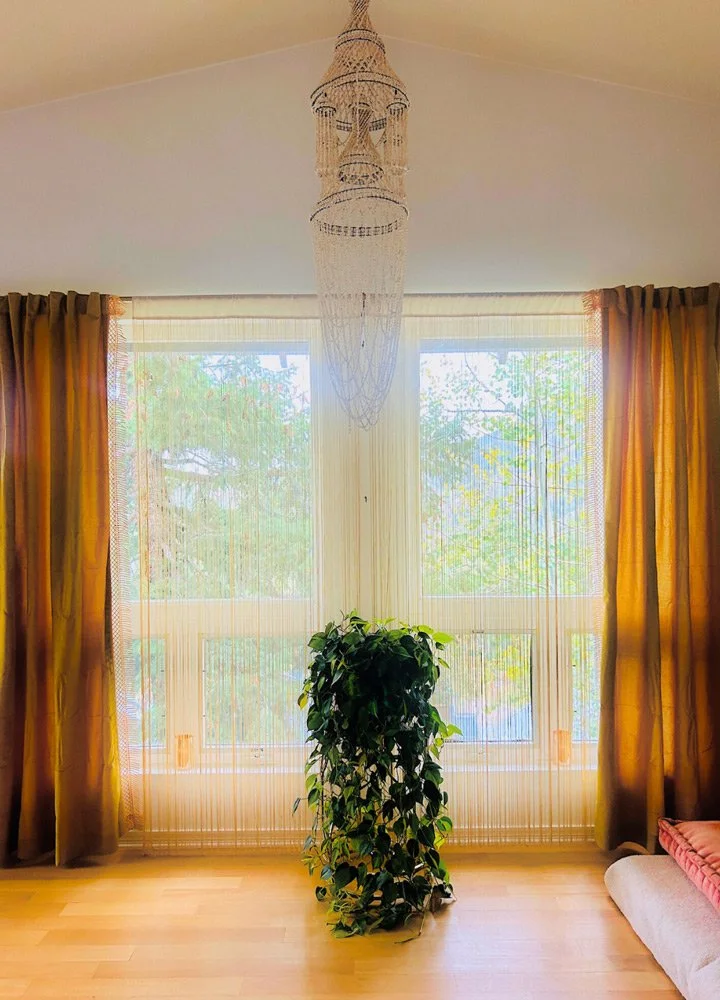 Bright living room with large window, yellow curtains, a green plant in the center, a chandelier hanging from the ceiling, wooden floor, and a pink cushion at Joytown, a transformational retreat & tantra sanctuary in Colorado.