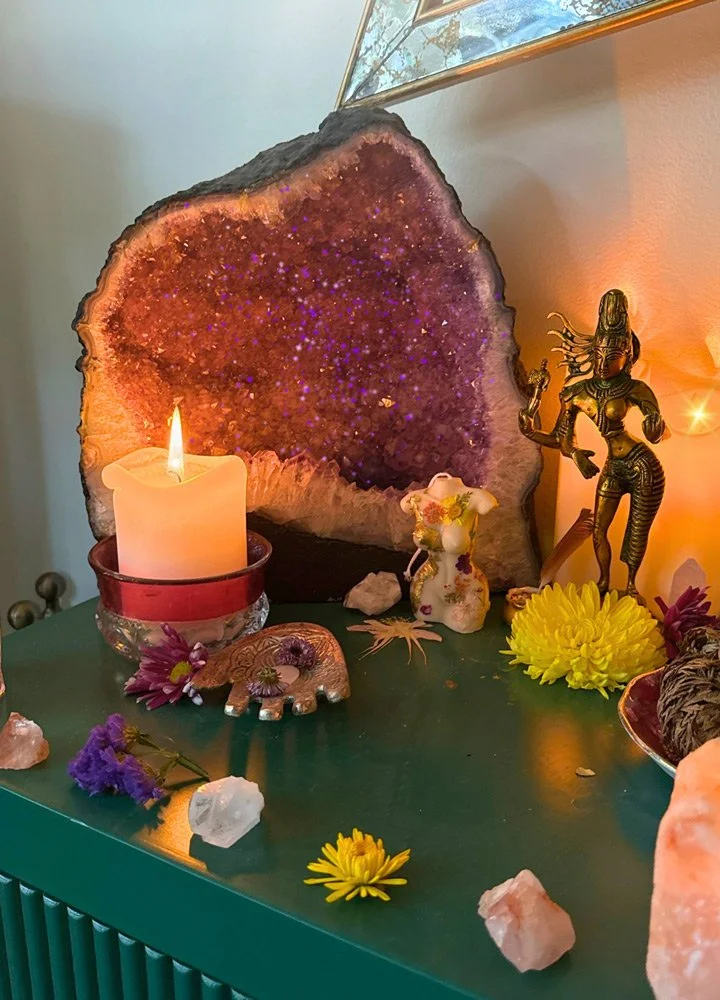 A decorative display on a green table featuring a large amethyst geode, a lit candle in a red holder, a small floral ceramic figure, and a bronze dancer statue at Joytown, a transformational retreat & tantra sanctuary in Colorado.