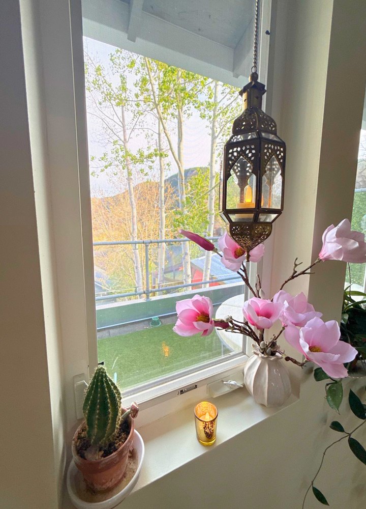 View from a window showing trees and a distant hill, with a decorative lantern hanging inside, next to a vase of pink flowers, a small cactus in a pot, and a lit candle on the window sill at Joytown, a transformational retreat & tantra sanctuary in C