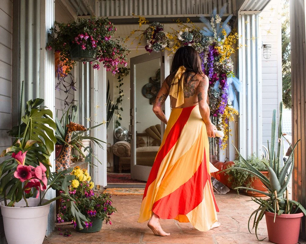 Woman with dark hair and tattoos wearing a colorful, flowing halter dress standing on a decorated outdoor patio with potted plants and a floral archway at Joytown, a transformational retreat & tantra sanctuary in Colorado.
