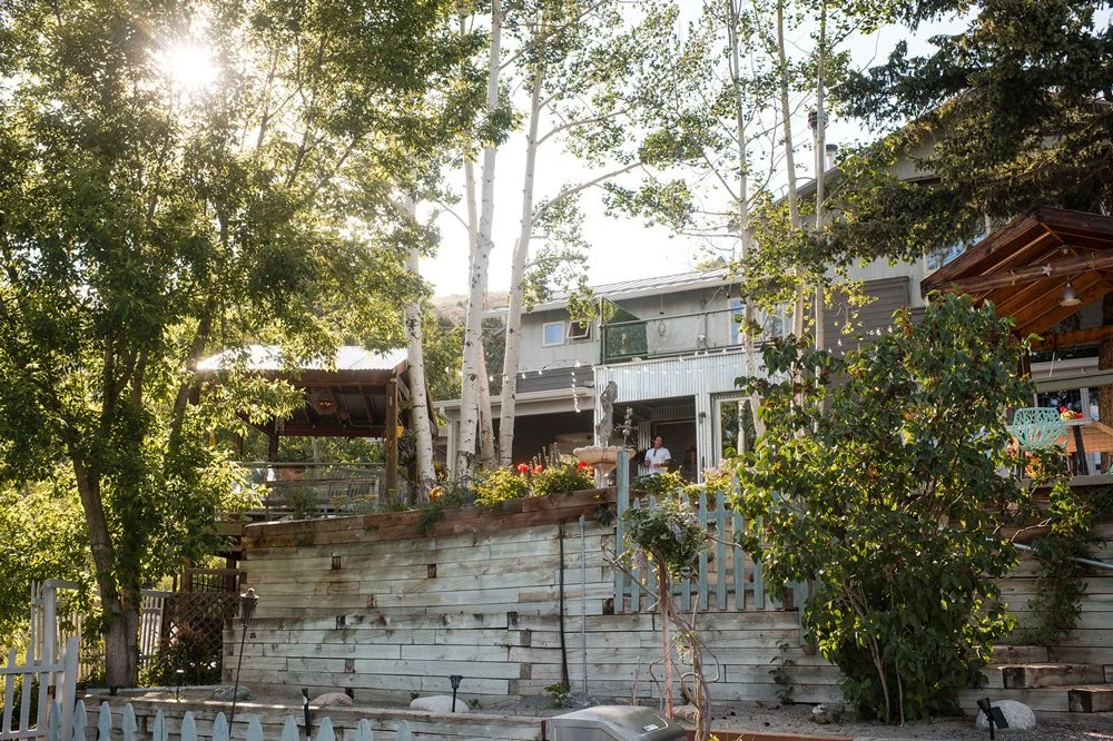 A backyard with a multi-level wooden deck, trees, and plants, with sunlight filtering through the leaves, and a person standing on the deck at Joytown, a transformational retreat & tantra sanctuary in Colorado.