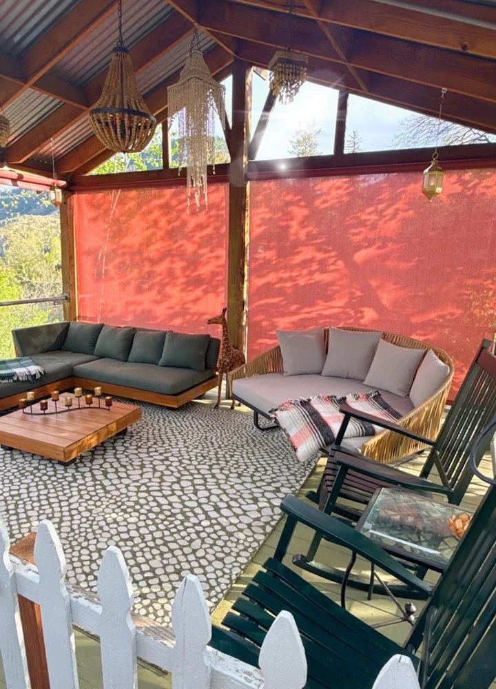A cozy outdoor porch with wicker and fabric sofas, a wooden coffee table with candles, hanging lanterns, chandeliers, a giraffe figurine, and a view of trees outside at Joytown, a transformational retreat & tantra sanctuary in Colorado.