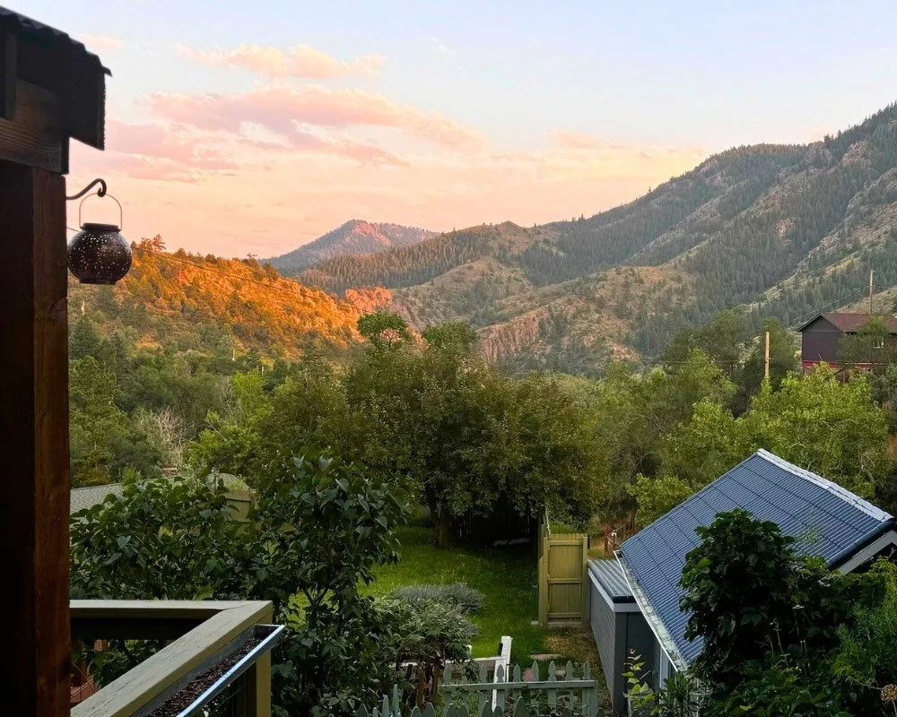 Scenic view of the mountains during sunset, with lush green trees, a blue-roofed shed, and parts of a wooden porch in the foreground at Joytown, a transformational retreat & tantra sanctuary in Colorado.