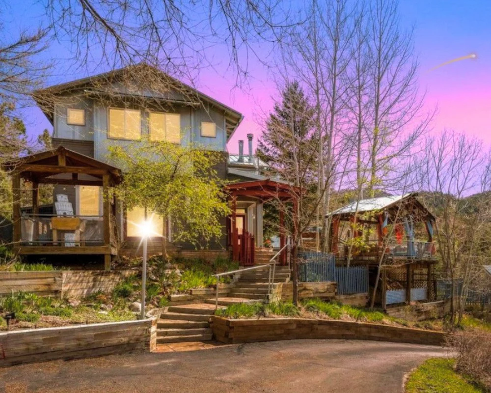 A two-story house with a porch and multiple windows, surrounded by trees and a garden with pathways, under a colorful sky at dusk or dawn at Joytown, a transformational retreat & tantra sanctuary in Colorado.