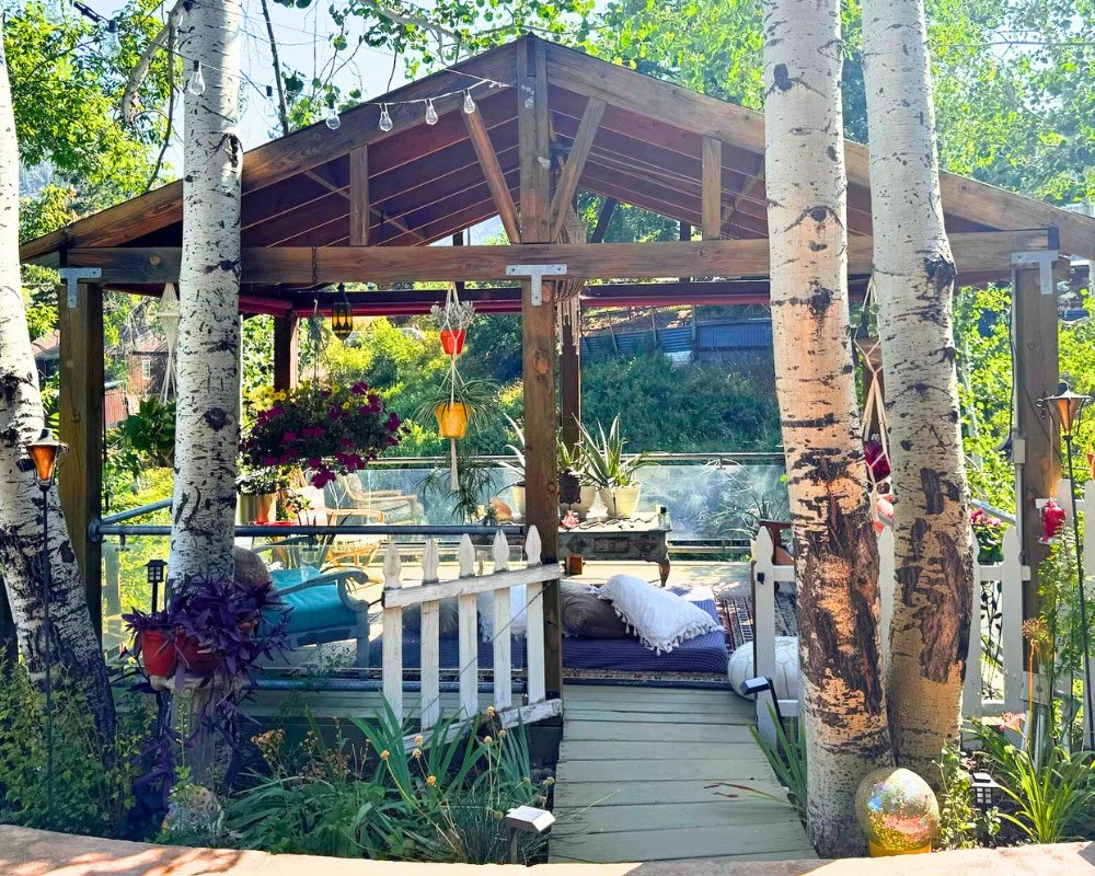 A cozy outdoor wooden porch with hanging lanterns and potted plants, surrounded by tall white birch trees, with garden and trees in the background at Joytown, a transformational retreat & tantra sanctuary in Colorado.
