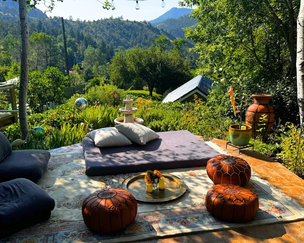 Outdoor seating area on a patterned rug with cushions and pillows, surrounded by lush greenery and overlooking mountains in the distance at Joytown, a transformational retreat & tantra sanctuary in Colorado.