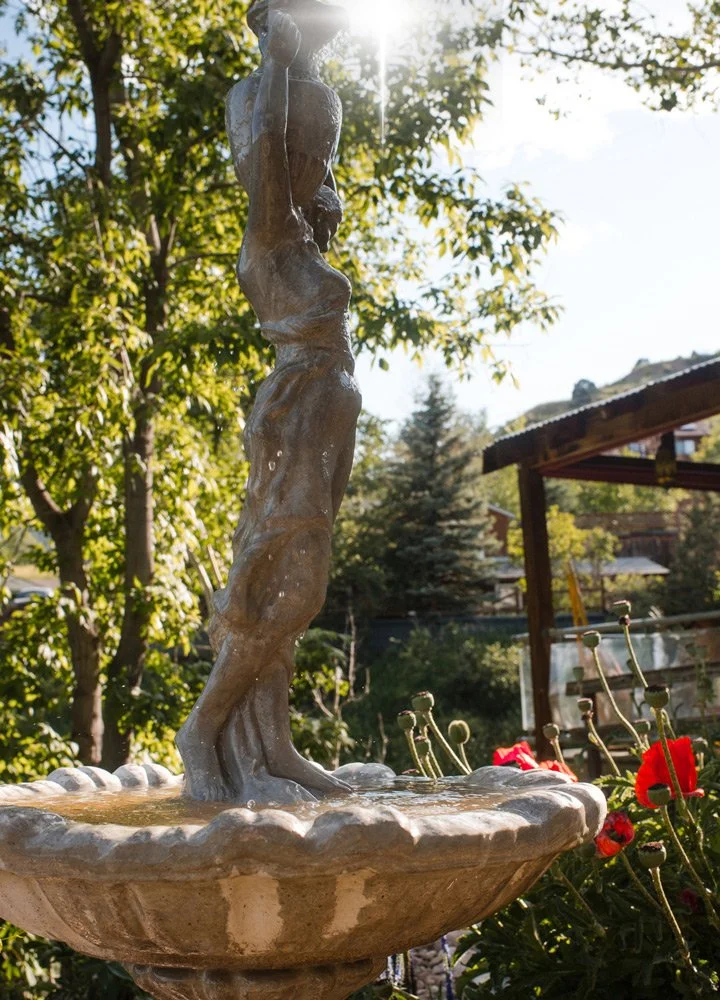 Outdoor stone fountain featuring a sculpture of a woman with water dripping from her hands, surrounded by greenery and flowers at Joytown, a transformational retreat & tantra sanctuary in Colorado.