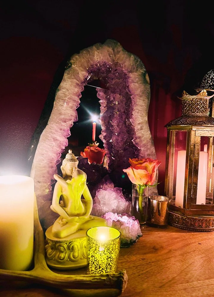 A decorative altar with a purple geode, a small Buddha statue, candles, and pink roses in a glass vase, set against a dark background at Joytown, a transformational retreat & tantra sanctuary in Colorado.